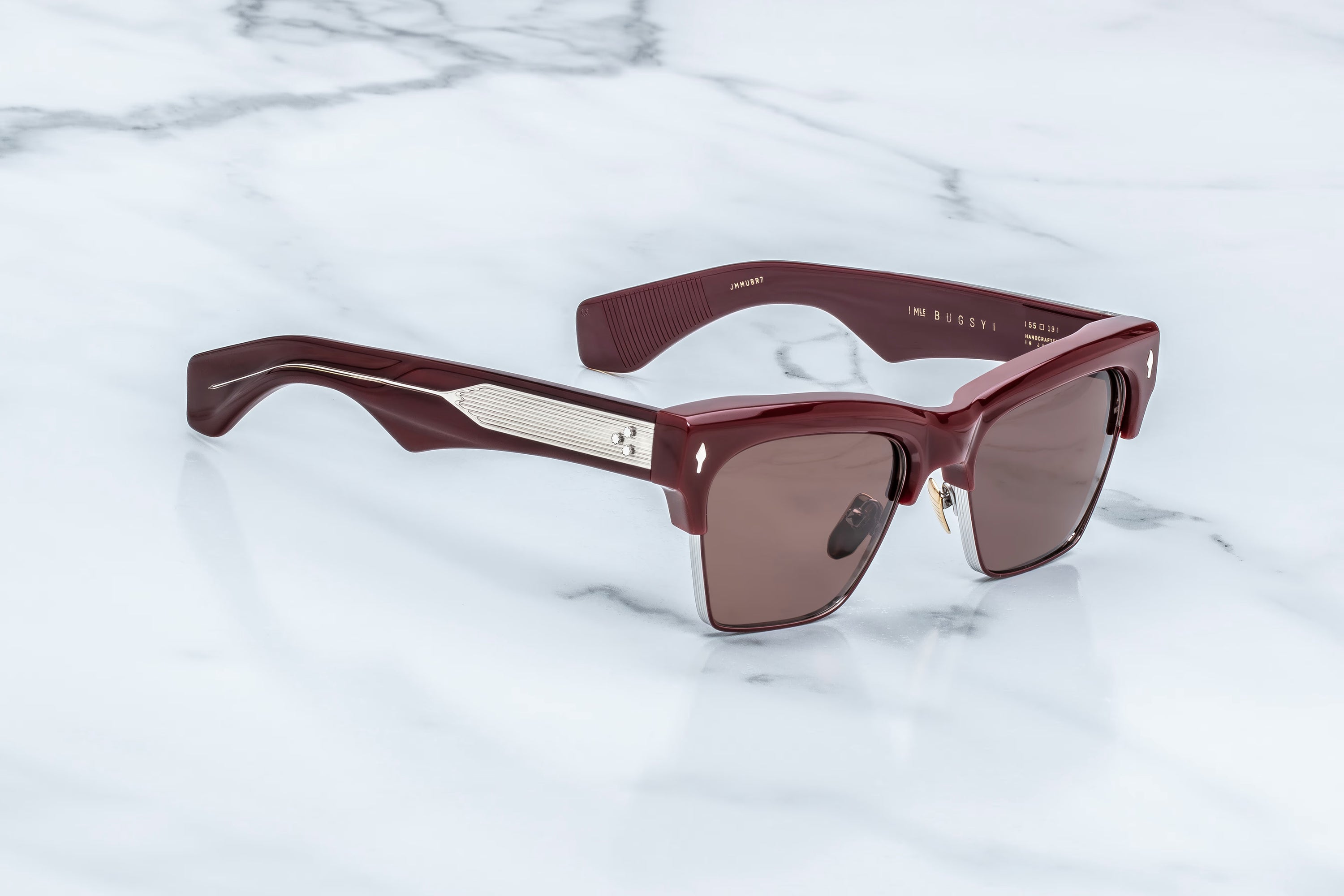 A pair of designer sunglasses with a glossy dark burgundy acetate frame, featuring a silver-tone metal accent on the front bridge and dark brown tinted lenses, resting on a white marble surface.