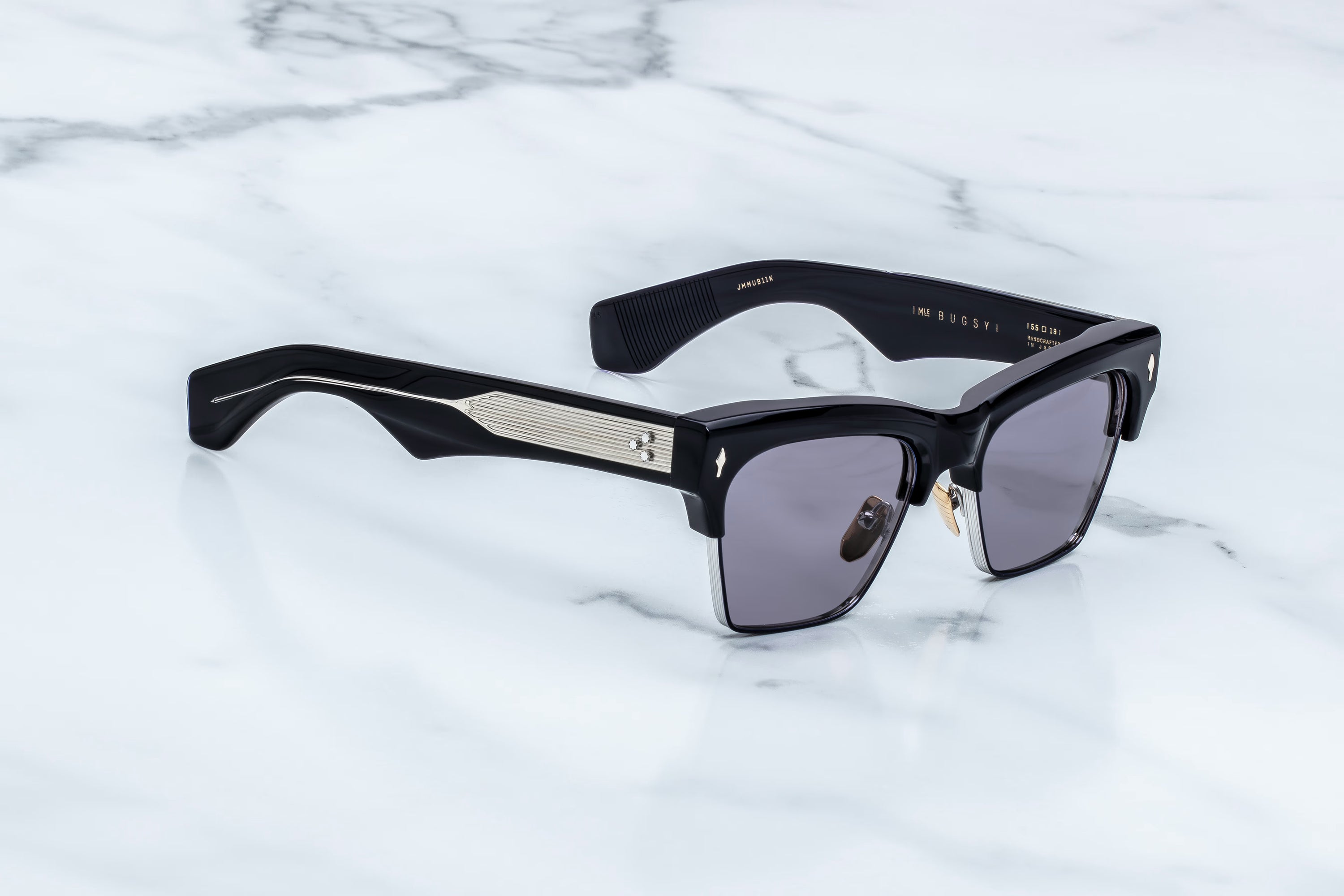 Black acetate sunglasses with dark gray lenses and silver metallic accents on the temples, displayed on a white marble surface.