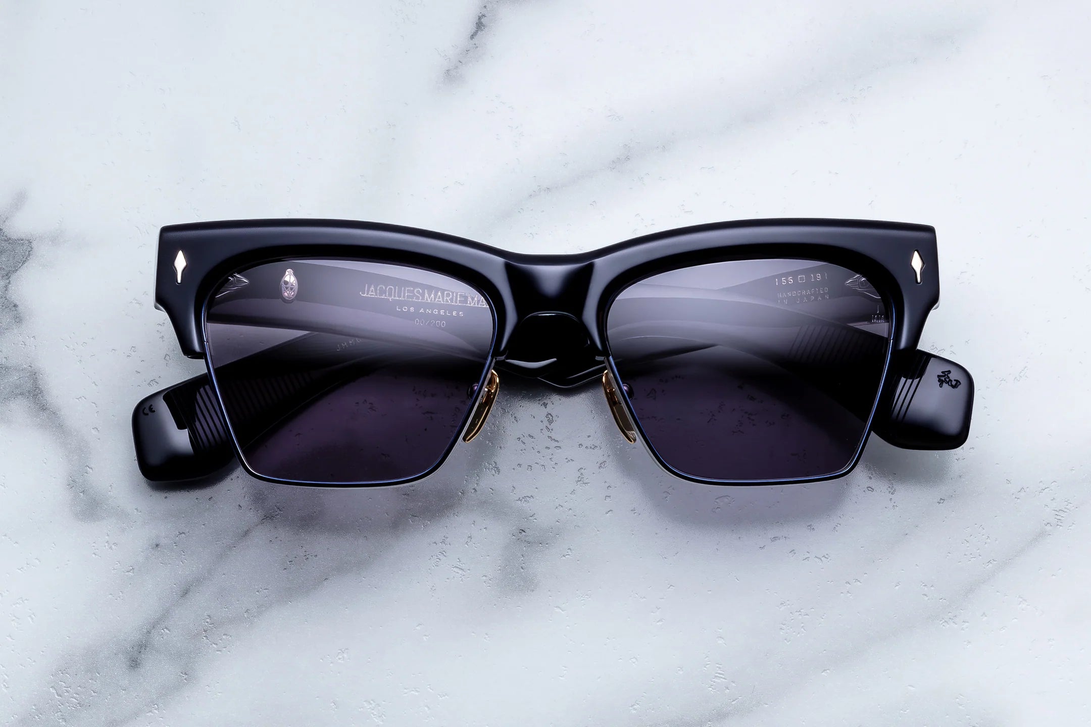 Black square sunglasses with dark tinted lenses and small gold-tone details, photographed from above on a white marble surface.