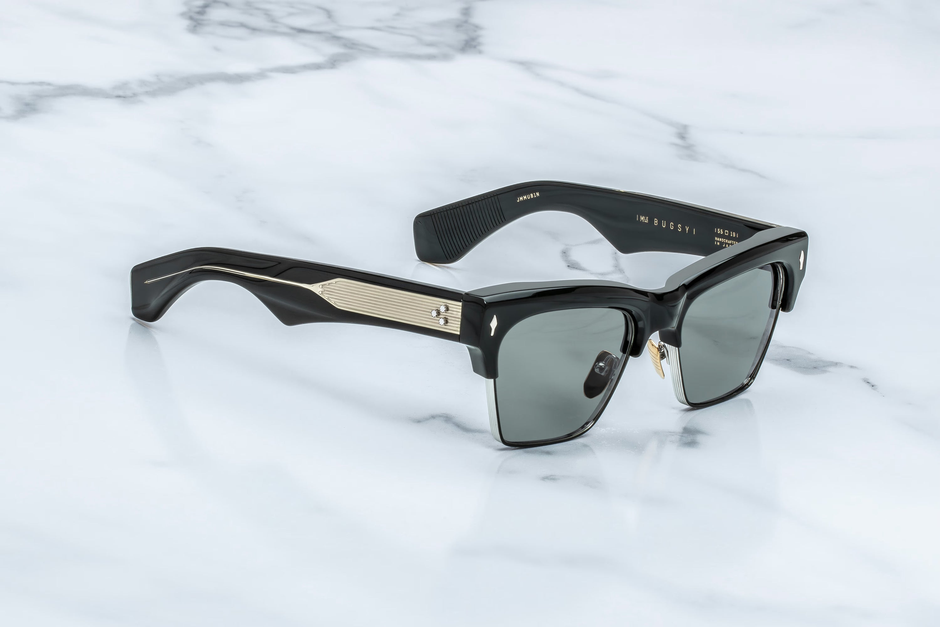 A pair of designer sunglasses with black acetate frames, gold-tone metal accents on the temples, and dark gray lenses, resting on a white marble surface.