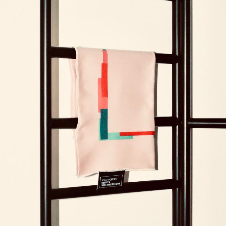 A pale blush pink silk scarf with a colorful geometric block pattern in red, teal, and orange, draped over a dark brown wooden ladder against a cream background.