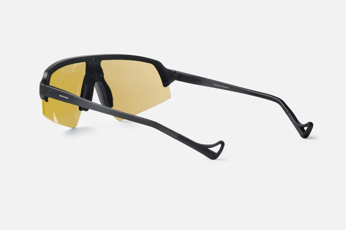 District Vision Choji Turbo Blade sunglasses by District Vision, featuring therapeutic-grade yellow-tinted lenses and a sleek black frame with curved arms, are displayed from an angled rear view against a plain light background—ideal for optimal eye protection during high-speed activities.