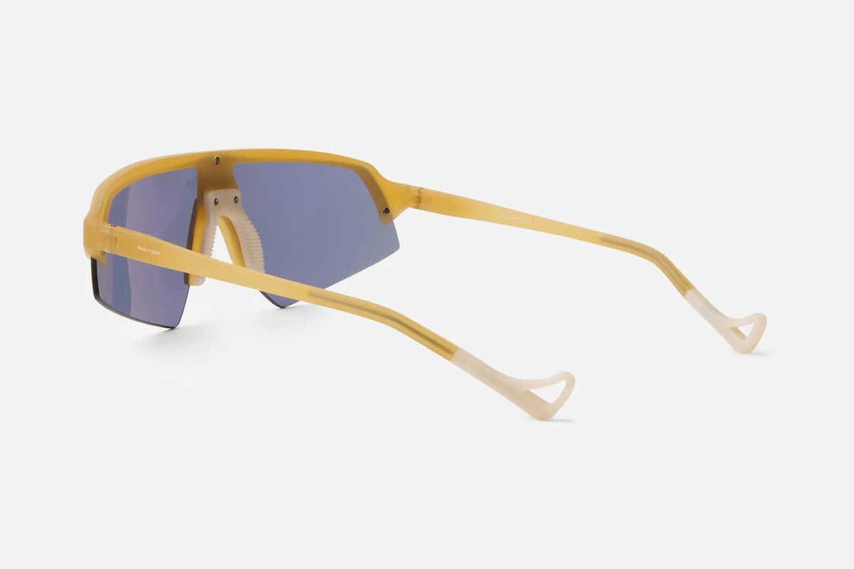 District Vision Choji Turbo Blade sunglasses by District Vision are displayed from an angled rear left view, highlighting their curved arms and modern geometric frame design in a vivid yellow finish with blue mirrored therapeutic-grade lenses, all set against a plain light background—perfect for stylish eye protection.