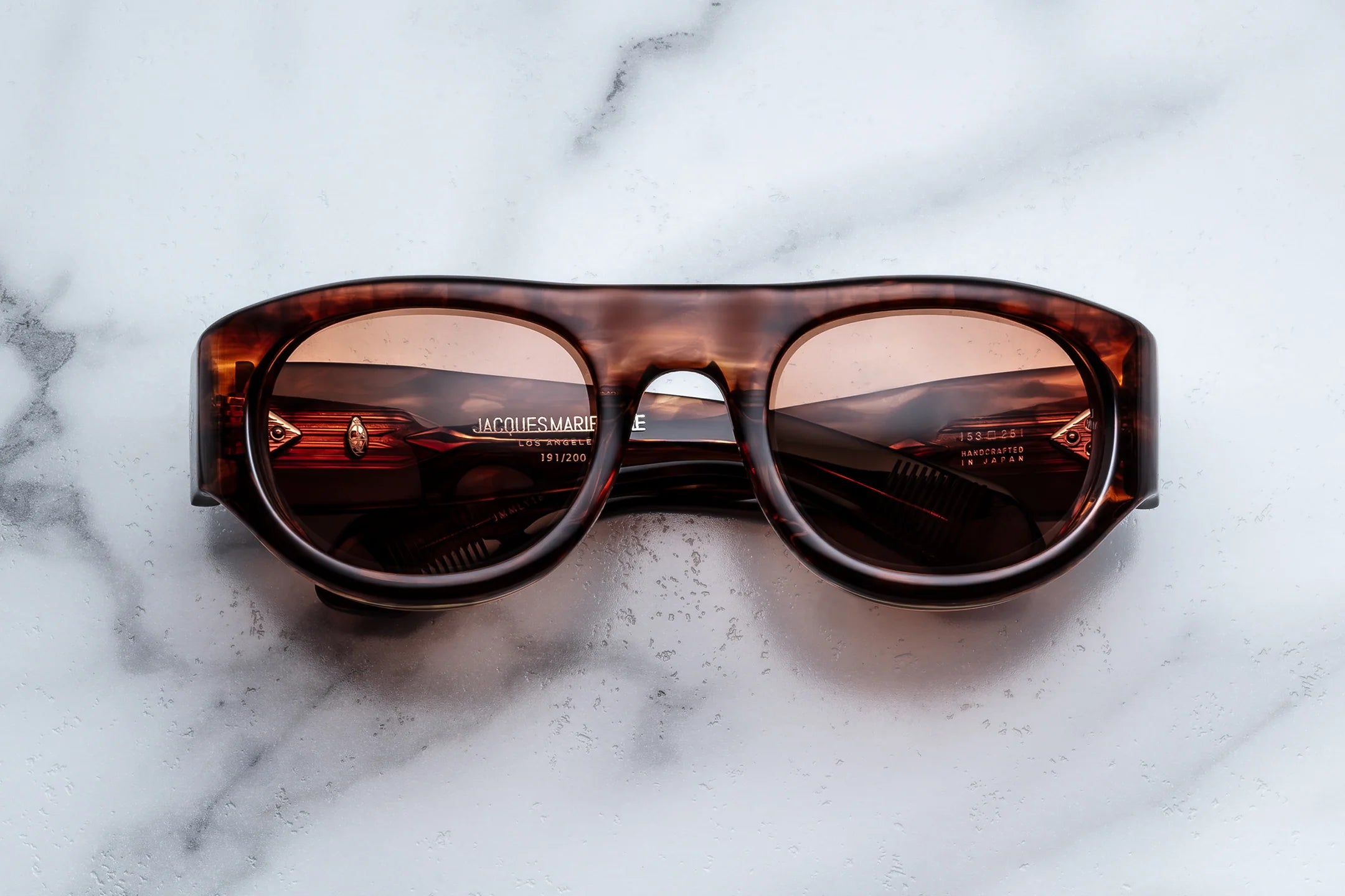 A pair of Jacques Marie Mage Clyde sunglasses in brown tortoiseshell, featuring thick frames, lies folded on a white marble surface, hinting at sophisticated wrap-around style and evoking a sense of freedom and fun, with the Jacques Marie Mage branding and product details visible on the inside of the arms.