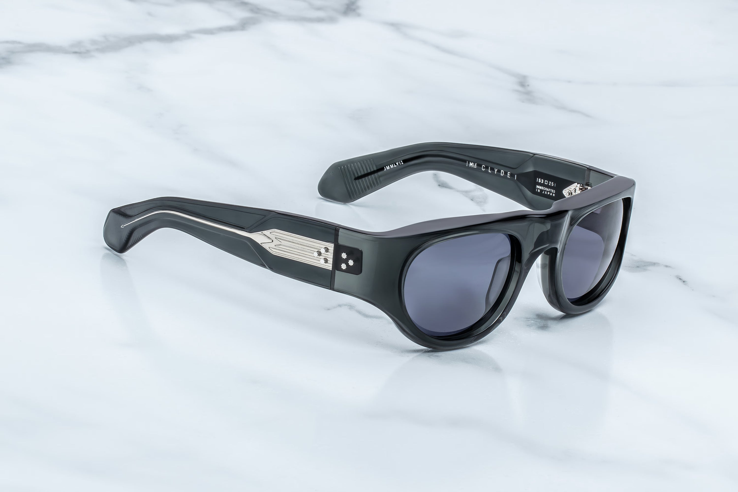 Jacques Marie Mage Clyde sunglasses by Jacques Marie Mage feature thick black frames and striking silver detail on the temple arms; these sophisticated wrap-around shades are displayed on a white marble surface, embodying a spirit of freedom and fun.