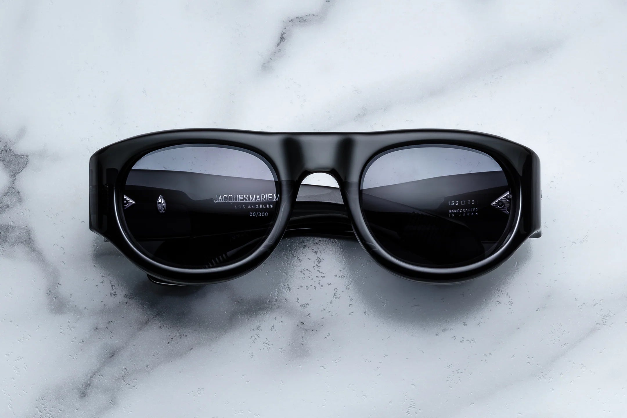 Jacques Marie Mage Clyde sunglasses with thick black frames are displayed face up on a white marble surface, their lenses catching visible reflections and the signature Jacques Marie Mage branding clearly marked on the inner arms. These sophisticated wrap-arounds blend impeccable style with practical functionality, ideal for anyone seeking freedom and fun in every sunny adventure.