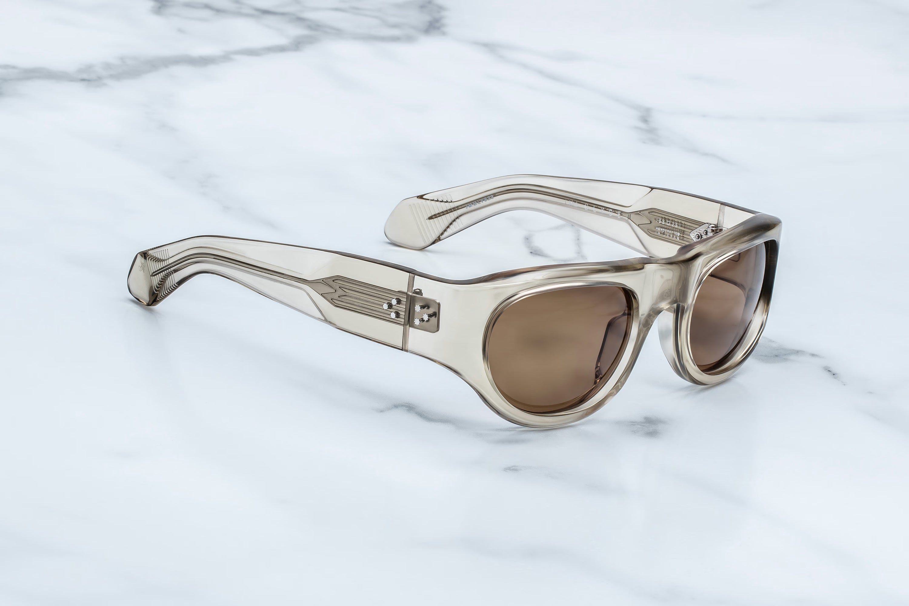The Jacques Marie Mage Clyde sunglasses by Jacques Marie Mage, featuring translucent beige thick frames and brown lenses, are displayed on a white marble surface, embodying a sense of freedom and fun in a chic, everyday hideaway.