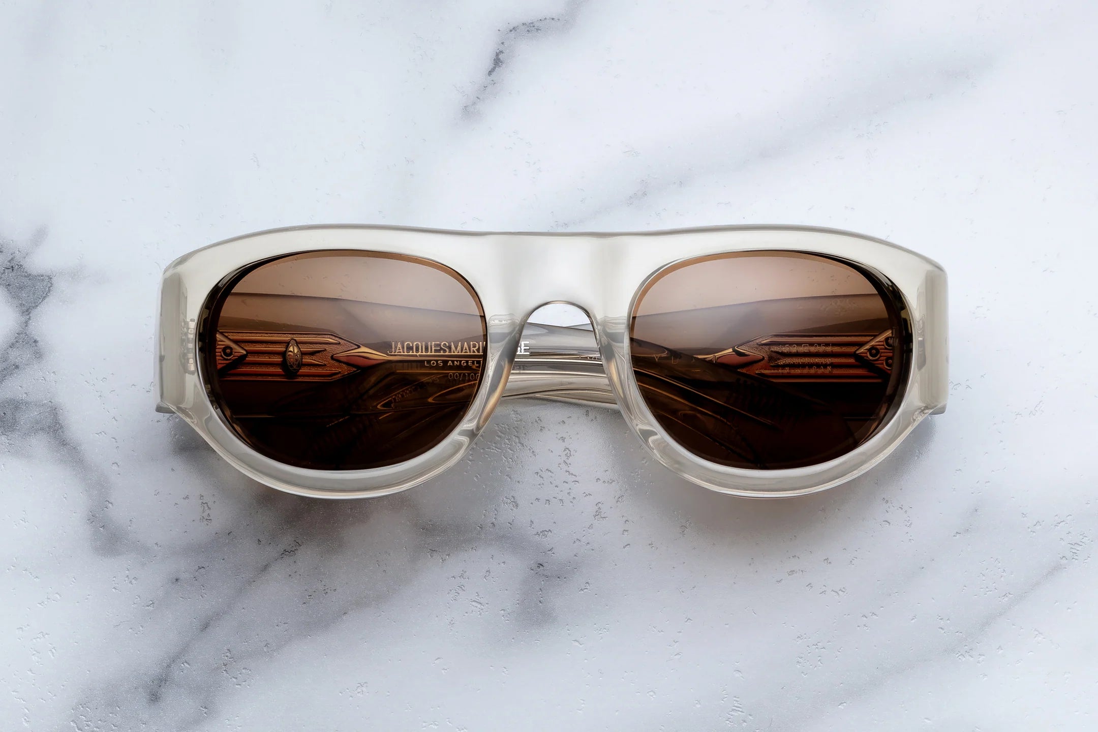 Jacques Marie Mage Clyde sunglasses featuring clear frames and brown-tinted lenses rest on a white marble surface. Evoking freedom and fun, these sophisticated wrap-arounds reflect metallic, angular objects and display a subtle Jacques Marie Mage logo on the left lens.