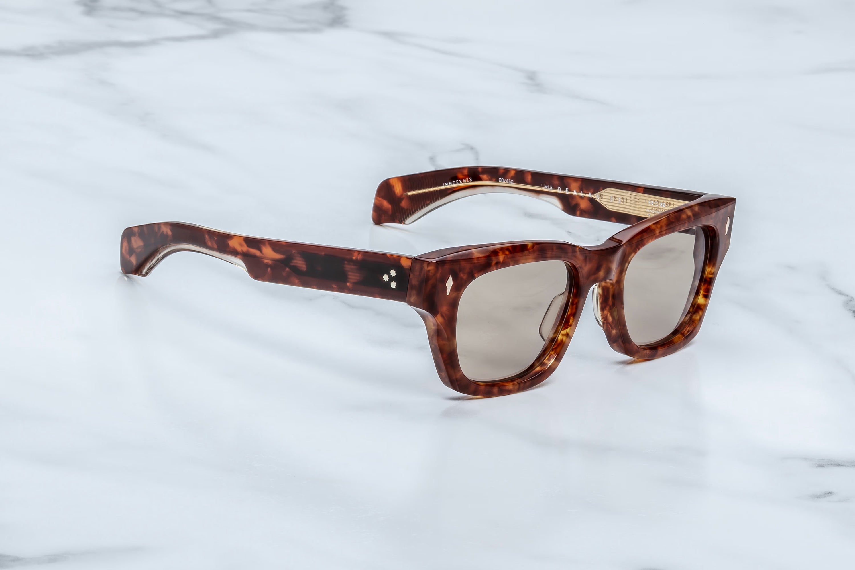 ’60s-inspired eyewear at its finest, the Jacques Marie Mage Dealan 53 sunglasses by Jacques Marie Mage feature a striking tortoiseshell square frame with brown lenses and are elegantly displayed on a white marble surface, showcasing timeless style.