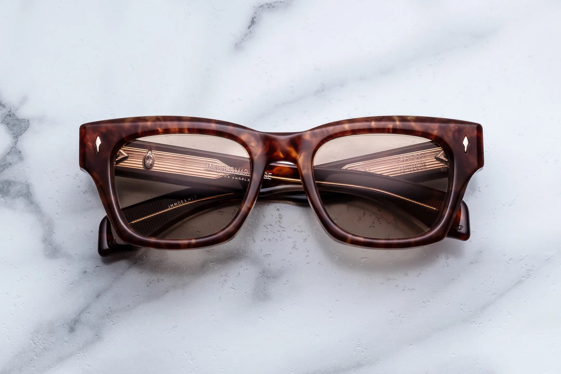 Jacques Marie Mage Dealan 53 sunglasses by Jacques Marie Mage, featuring a brown tortoiseshell rectangular frame with dark lenses inspired by ’60s eyewear, are displayed on a light marble surface. The arms of the sunglasses are folded, showcasing intricate detailing inside the frames and reflecting their timeless style.