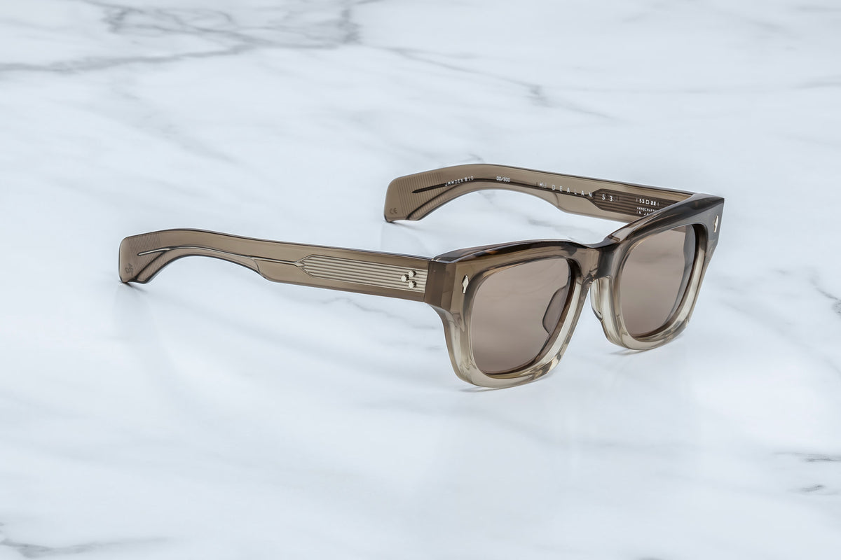 The Jacques Marie Mage Dealan 53 by Jacques Marie Mage, featuring brown-tinted rectangular lenses and thick frames inspired by 60s-era eyewear, rests on a white marble surface.