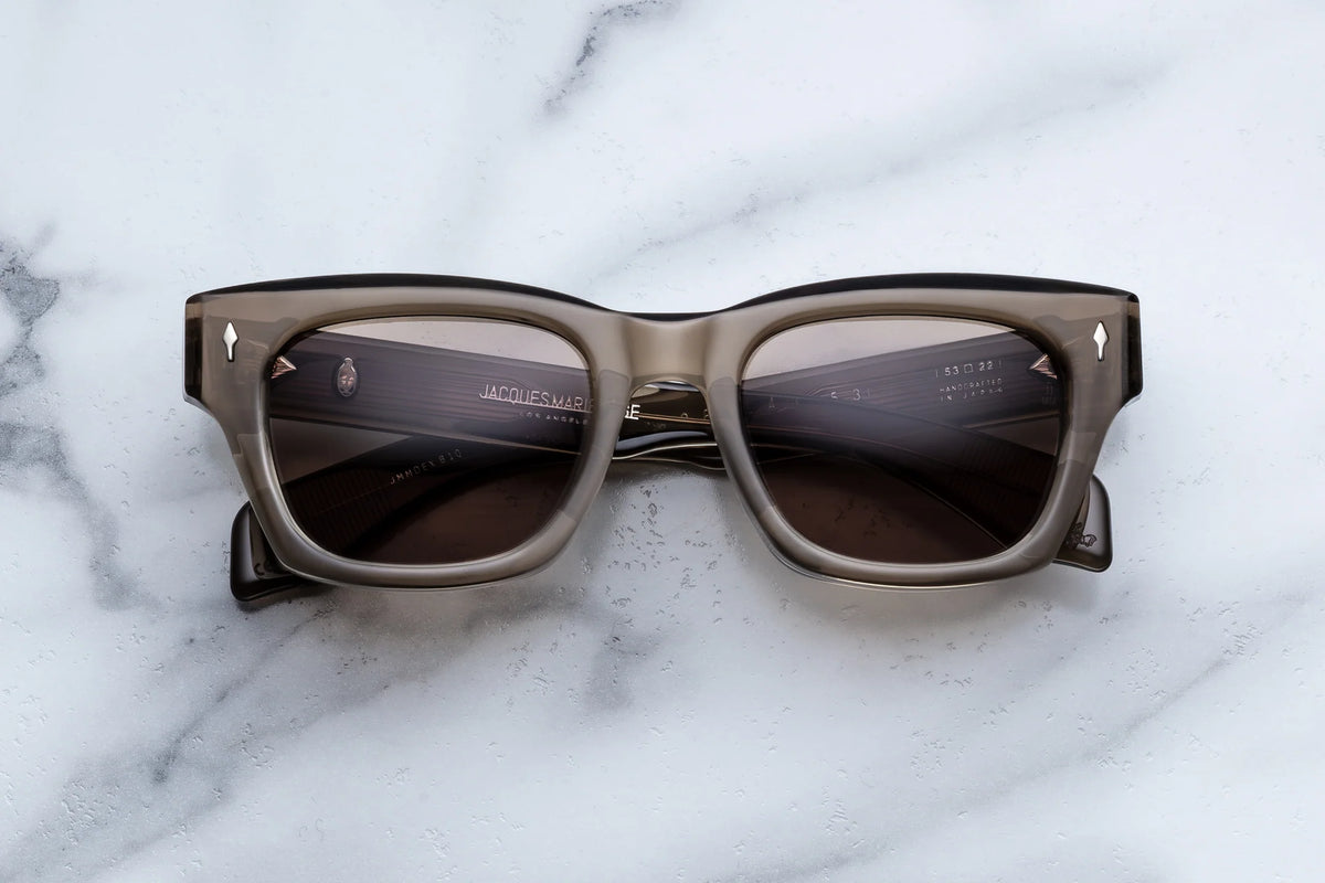 Rectangular, thick-framed gray Jacques Marie Mage Dealan 53 sunglasses with dark lenses rest on a white marble surface, embodying the timeless style of 60’s-inspired eyewear from Jacques Marie Mage.