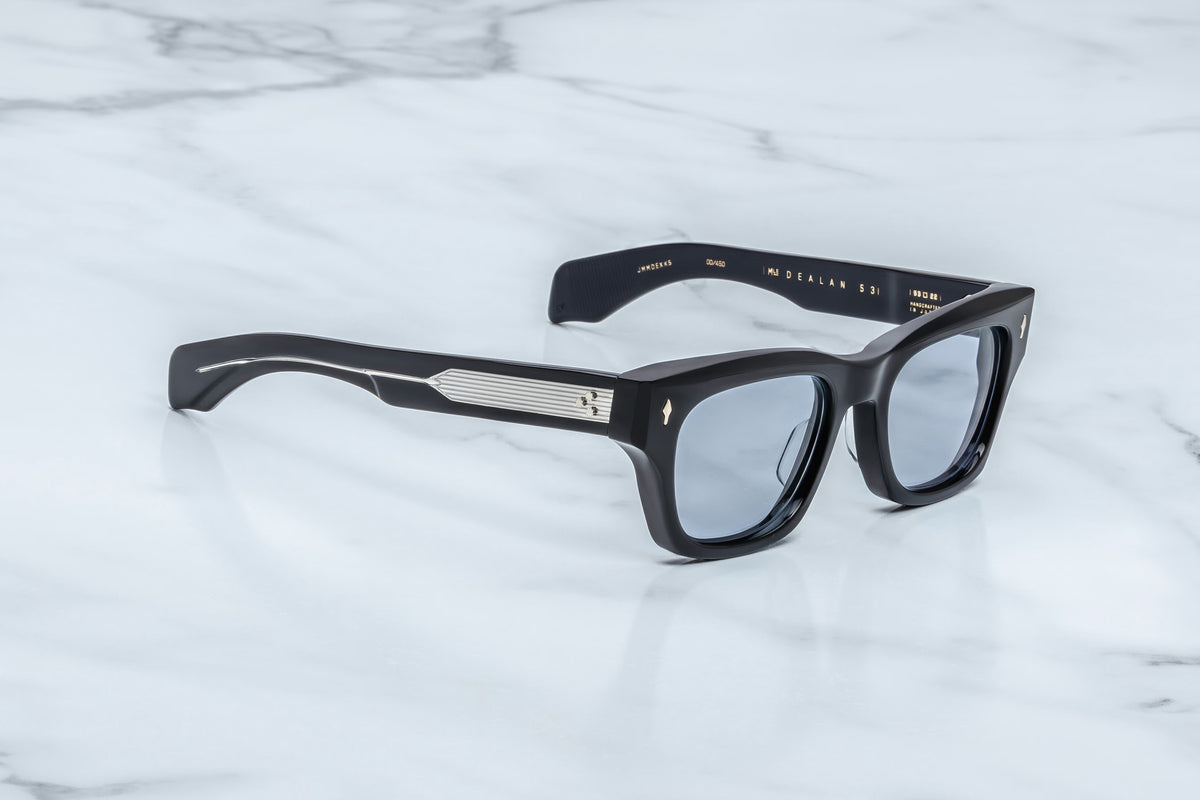 The Jacques Marie Mage Dealan 53 by Jacques Marie Mage showcases black rectangular sunglasses with thick frames and silver accents on the temples, displayed on a white marble surface. Inspired by 60s eyewear, this pair offers a timeless style perfect for any occasion.