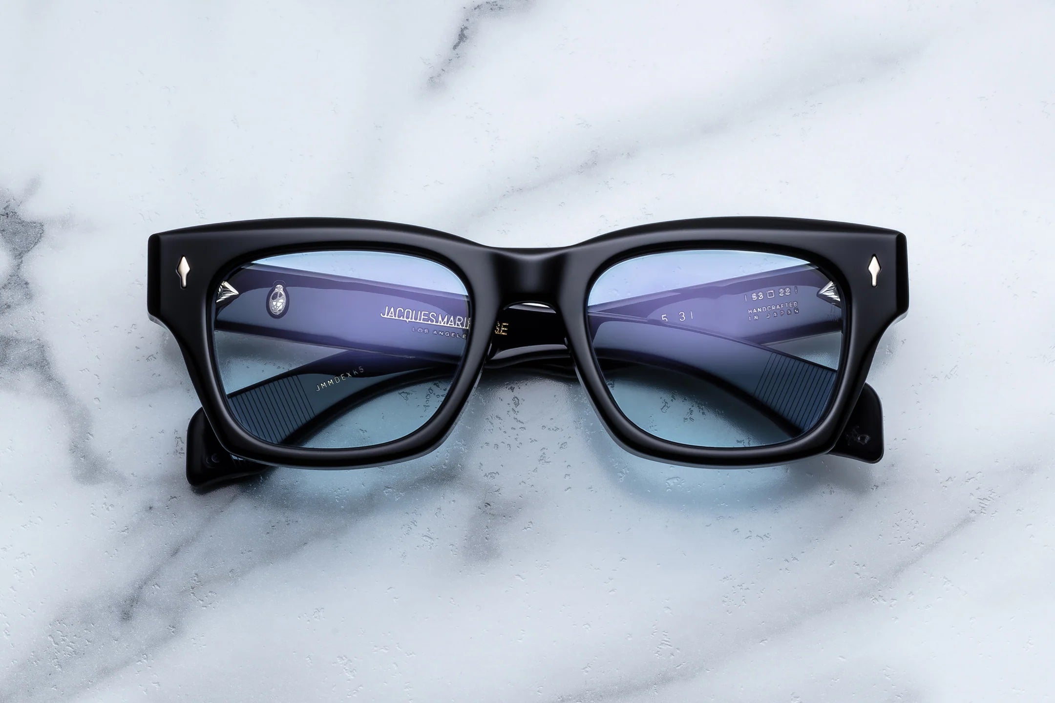 Jacques Marie Mage Dealan 53 eyeglasses by Jacques Marie Mage, featuring a bold black frame and blue-tinted lenses, rest on a white marble surface. Inspired by iconic 60s eyewear, these glasses showcase visible branding on the inner arms and silver accents at the front corners, embodying timeless style.