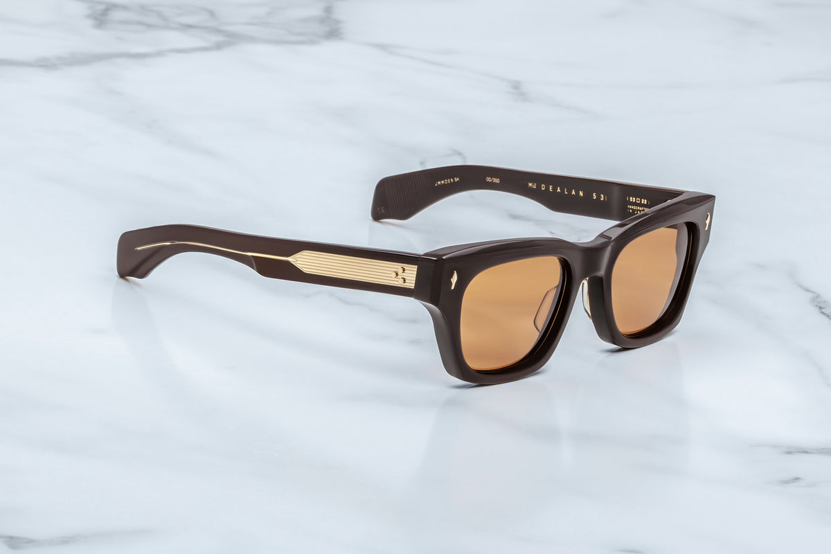 The Jacques Marie Mage Dealan 53 sunglasses by Jacques Marie Mage feature brown tinted lenses set within thick frames accented with gold detailing, resting elegantly on a white marble surface. Their bold, modern silhouette captures the spirit of 60’s-inspired eyewear, delivering a truly timeless style.