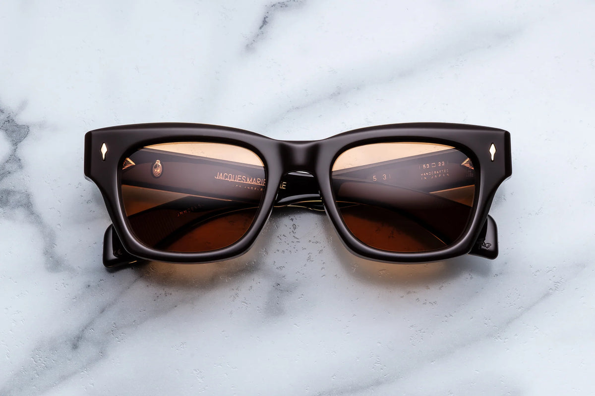 The Jacques Marie Mage Dealan 53 sunglasses by Jacques Marie Mage, featuring black rectangular frames with brown lenses, are displayed on a white marble surface. Part of the renowned THE DEALAN collection, these sunglasses showcase thick frames and subtle metallic accents at the corners, embodying 60s-inspired design and timeless sophistication.
