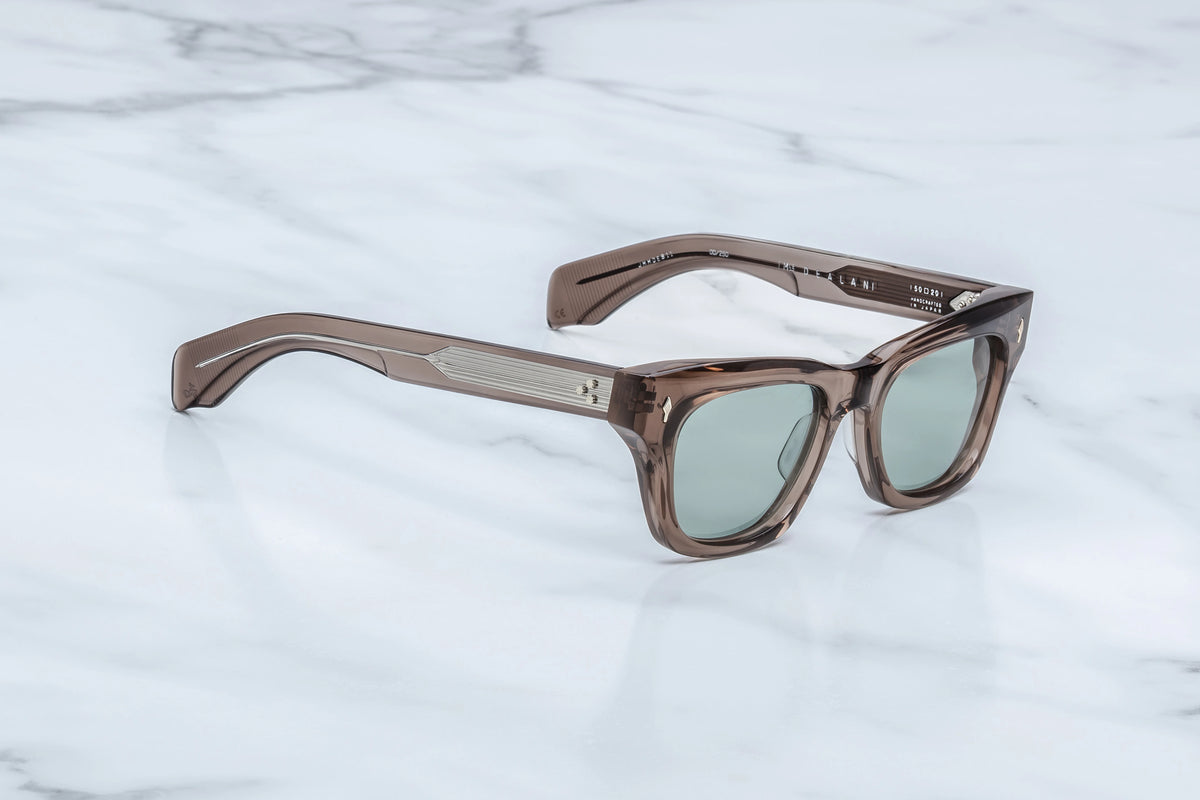 Translucent brown rectangular Jacques Marie Mage Dealan sunglasses with light green lenses resting on a white marble surface.