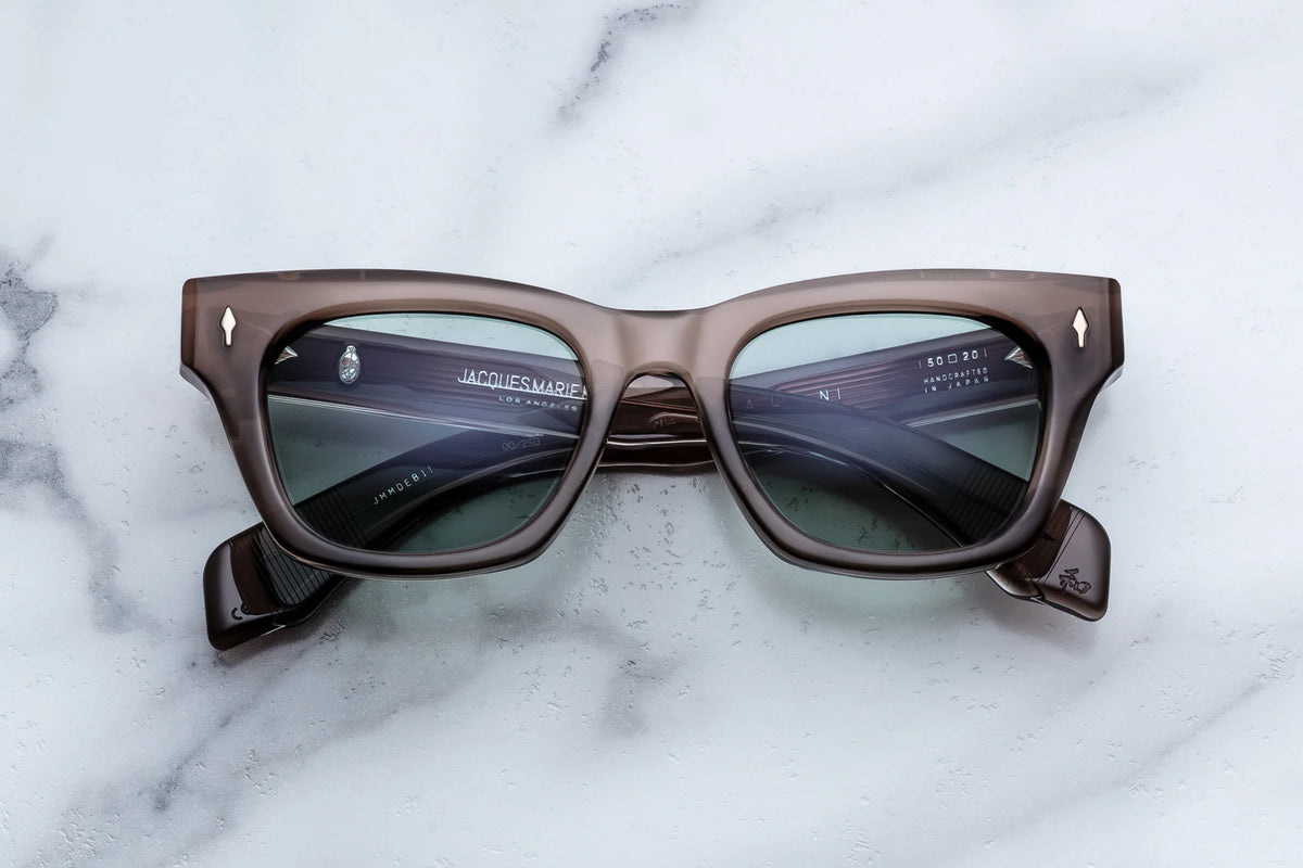 Translucent brown rectangular Jacques Marie Mage Dealan sunglasses with grey-tinted lenses resting on a marble surface, capturing a sophisticated ‘60s aesthetic.