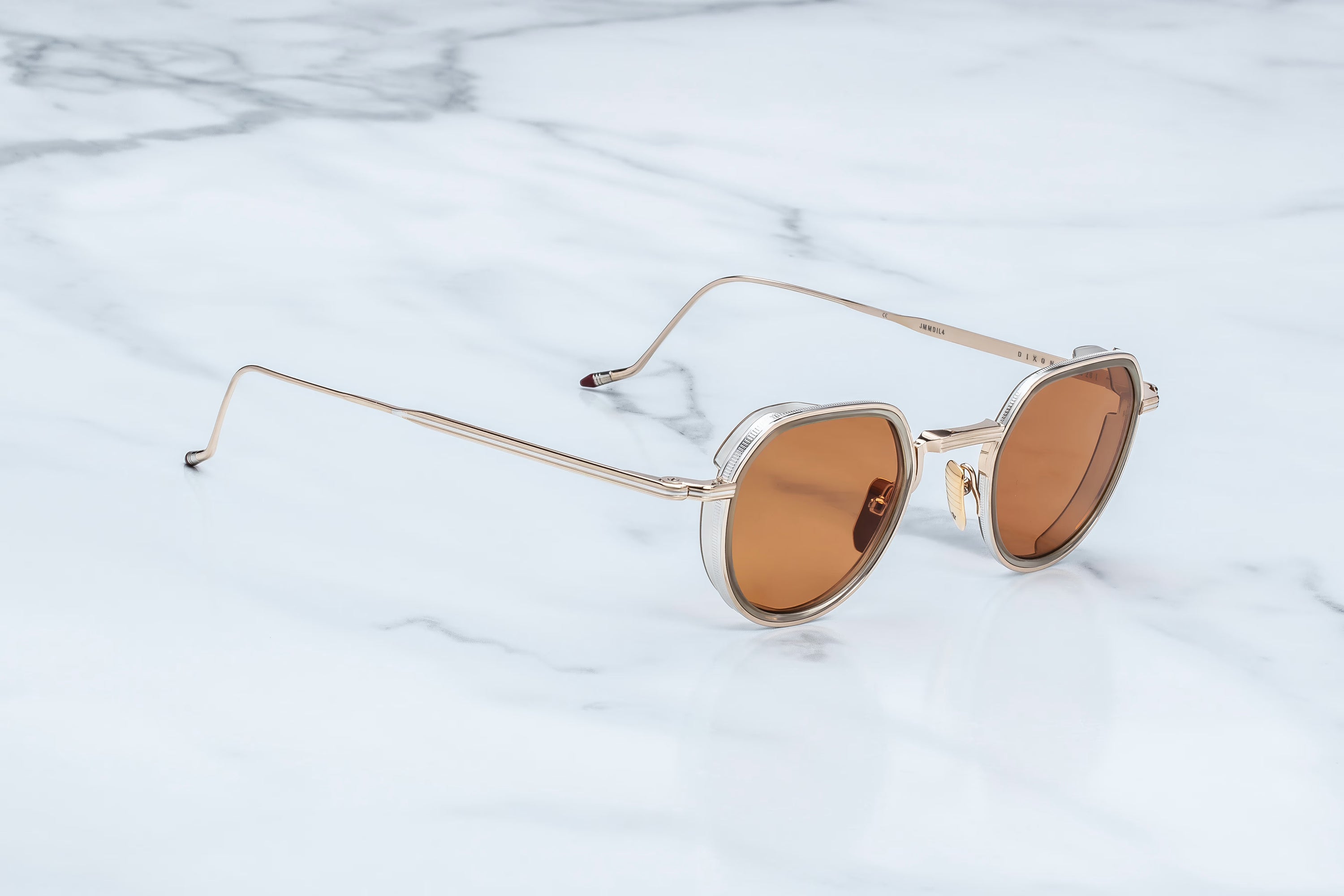 Oval sunglasses with a gold-tone metal frame and warm brown lenses, resting on a white marble surface.