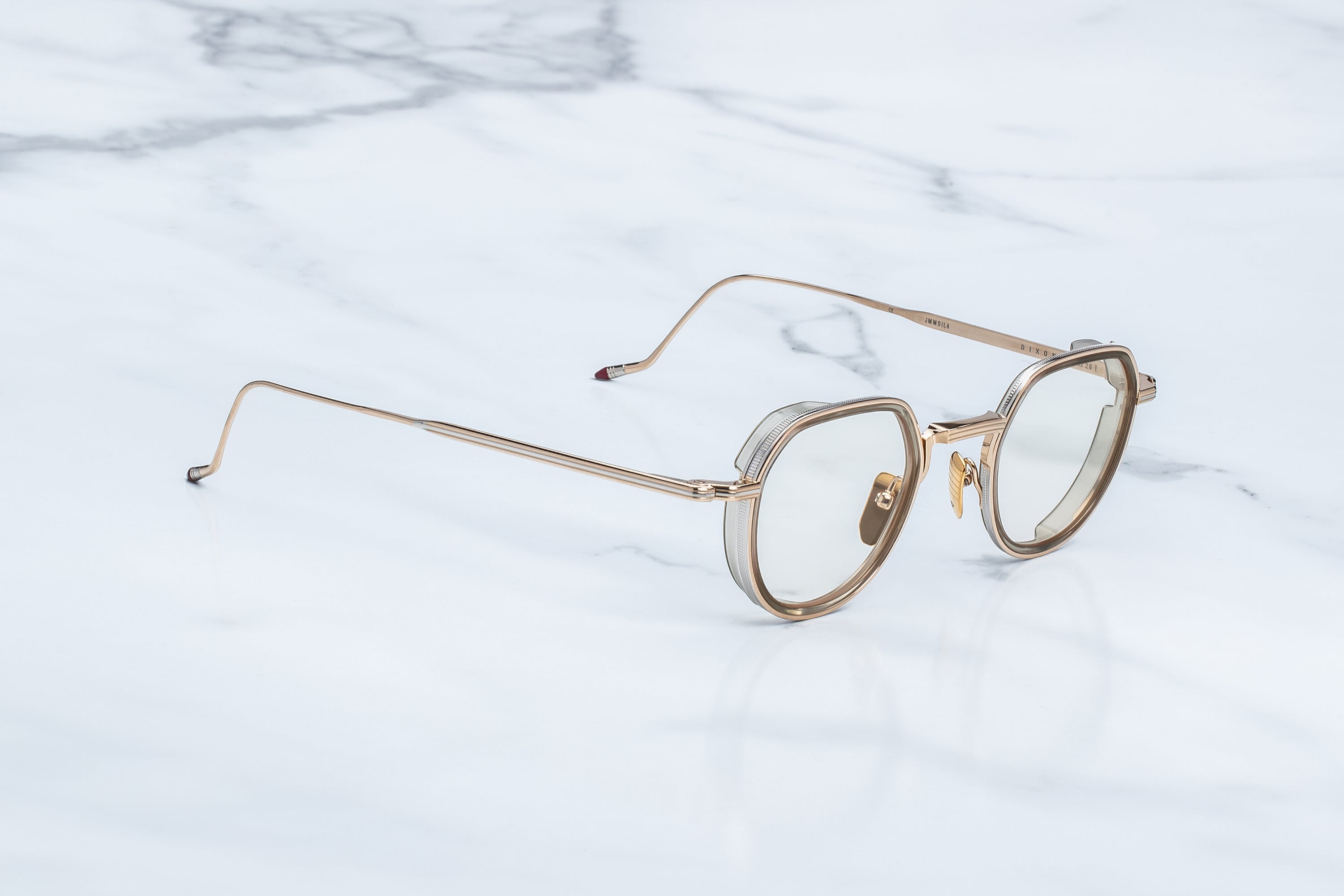 Jacques Marie Mage Dixon eyeglasses by Jacques Marie Mage, with their gold frames and round, clear lenses, rest on a white marble surface—evoking the quiet intellect of rogue philosophers and the unmistakable outlaw spirit of those who see the world differently.