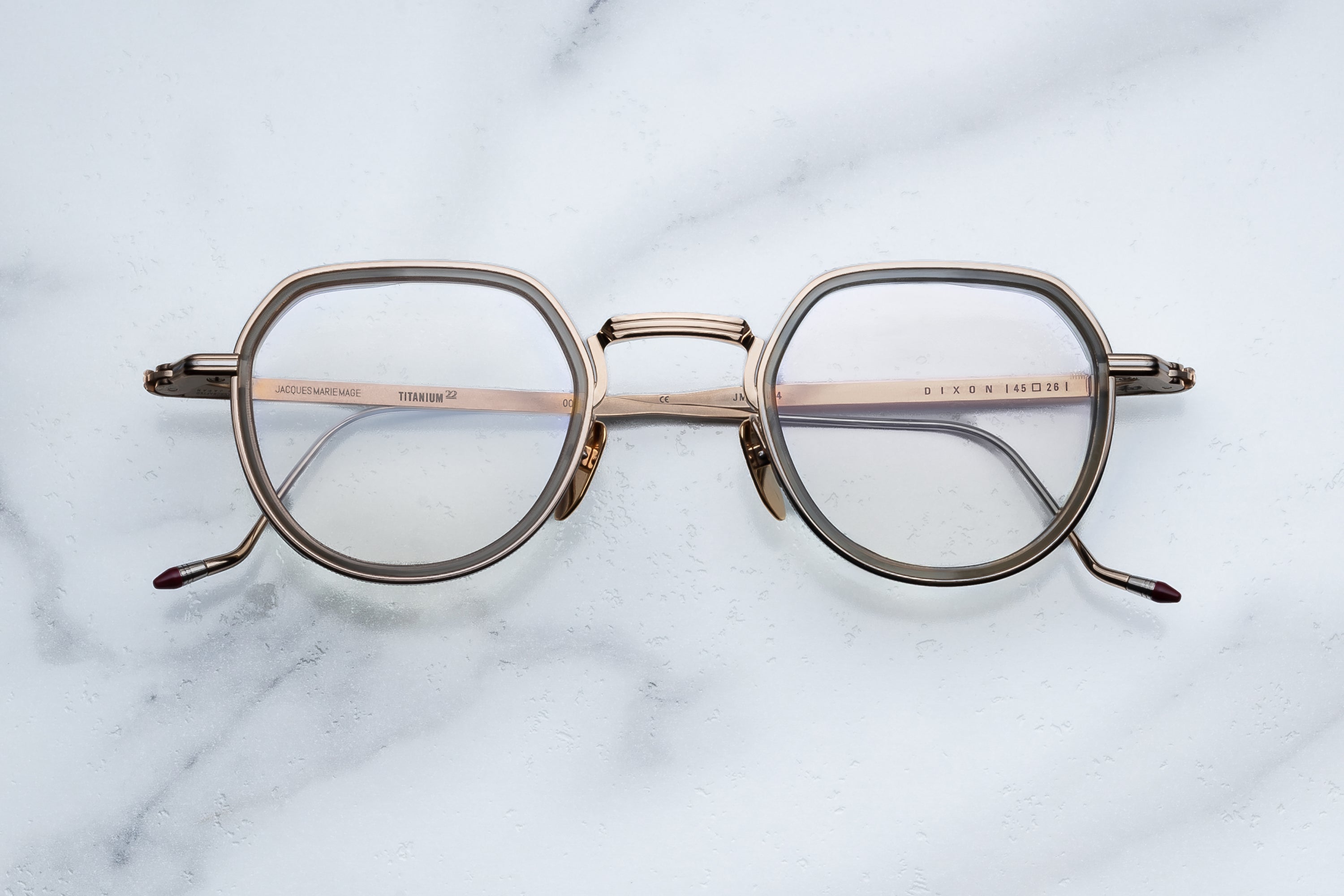 A pair of Jacques Marie Mage Dixon gold metal eyeglasses with clear lenses rests on a white marble surface. The geometric frame, featuring thin arms and a subtle bridge design, captures the refined edge of rogue philosophers—ideal for those who value clarity paired with the outlaw spirit of Jacques Marie Mage craftsmanship.