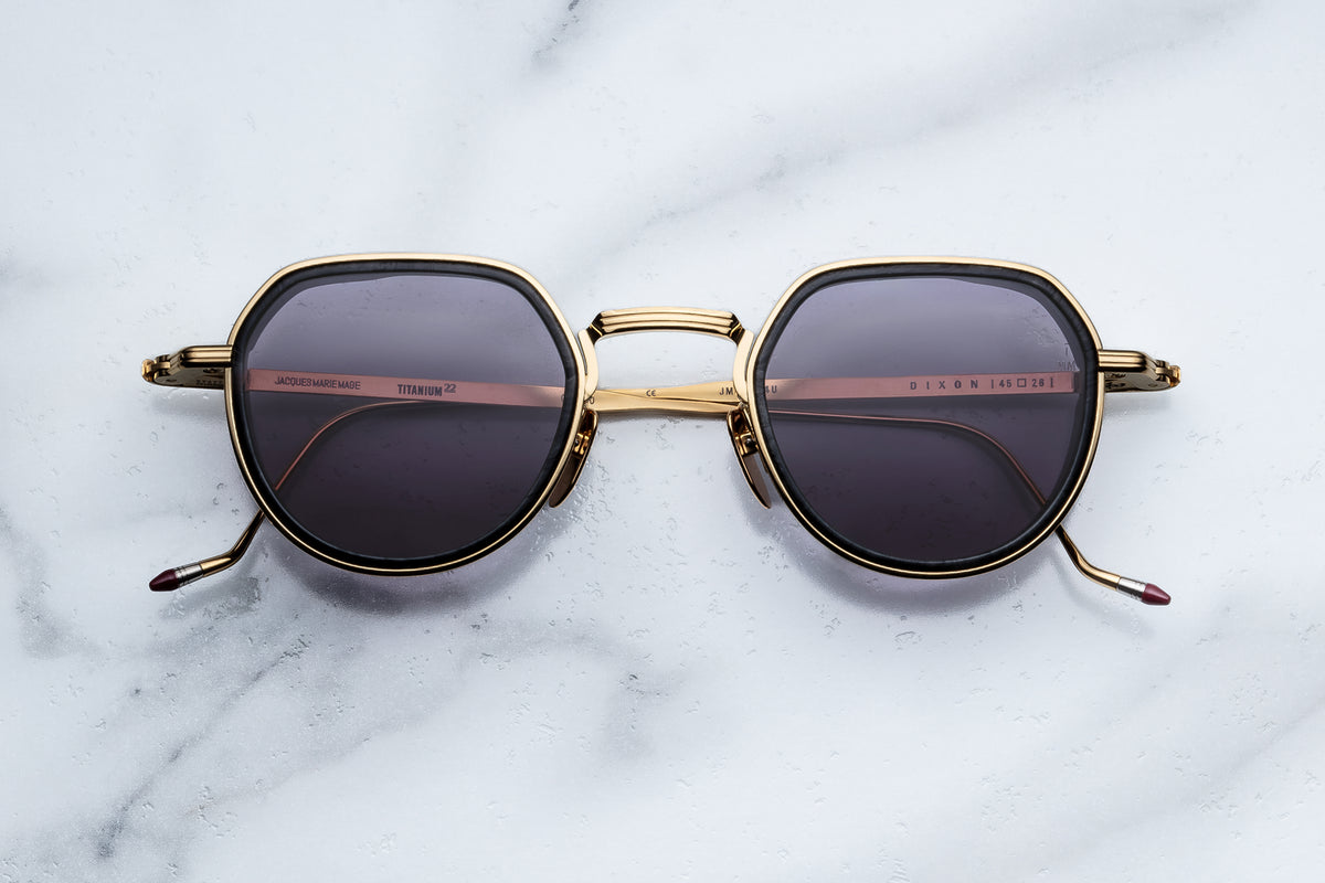 A pair of gold-framed round sunglasses with dark lenses, resting on a white marble surface.