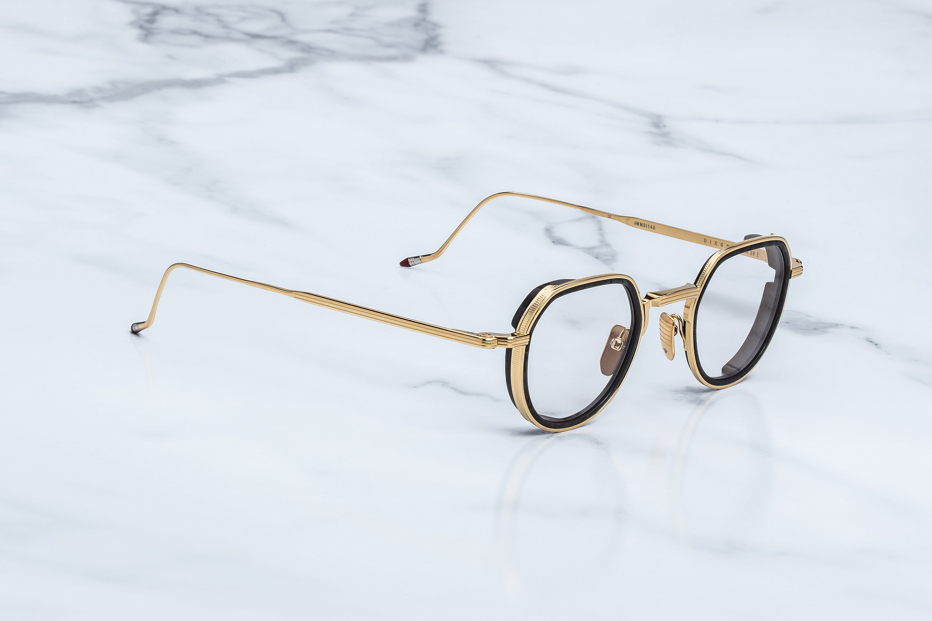Jacques Marie Mage Dixon eyeglasses by Jacques Marie Mage—featuring gold-rimmed round frames with striking black accents—rest on a white marble surface, viewed from a gentle angle; an accessory destined for rogue philosophers and anyone with an outlaw spirit.