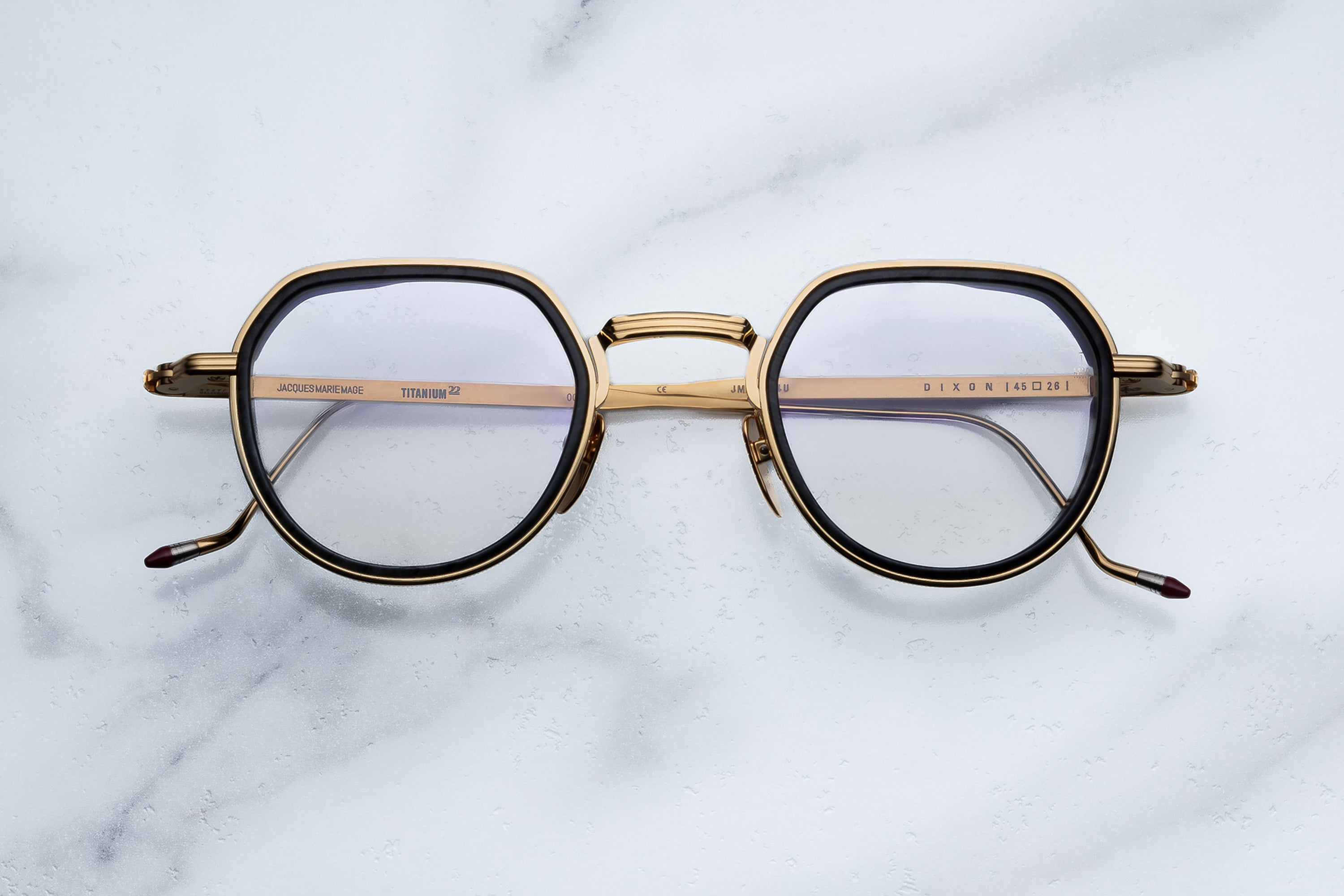 Jacques Marie Mage Dixon eyeglasses by Jacques Marie Mage rest atop a white marble surface, viewed from above. These gold, round frames feature thin metallic construction and dark brown accents on the temple tips, channeling an outlaw spirit that blends timeless elegance with subtle rebellion.