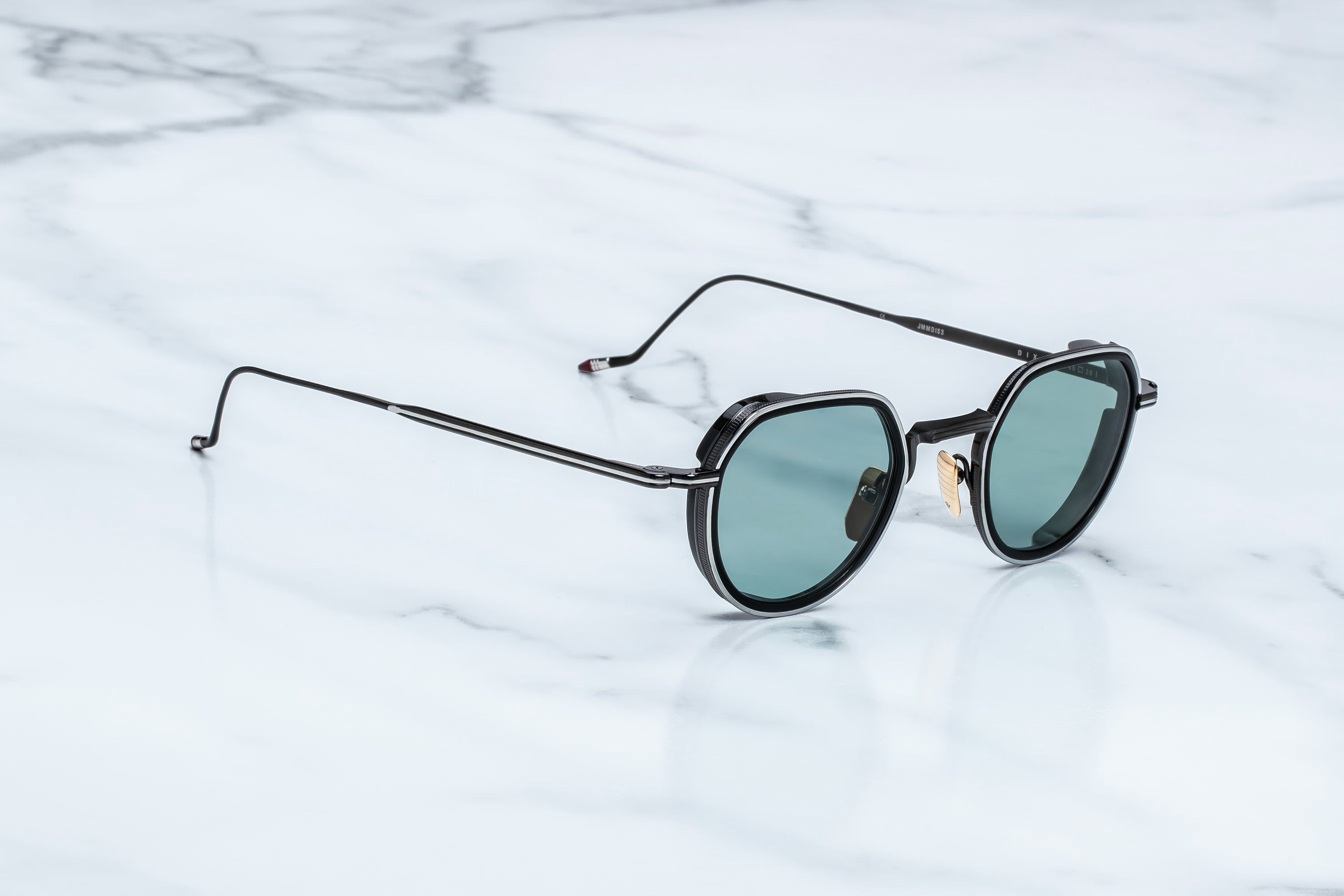 Round sunglasses with a black metal frame and green-tinted lenses, resting on a white marble surface.