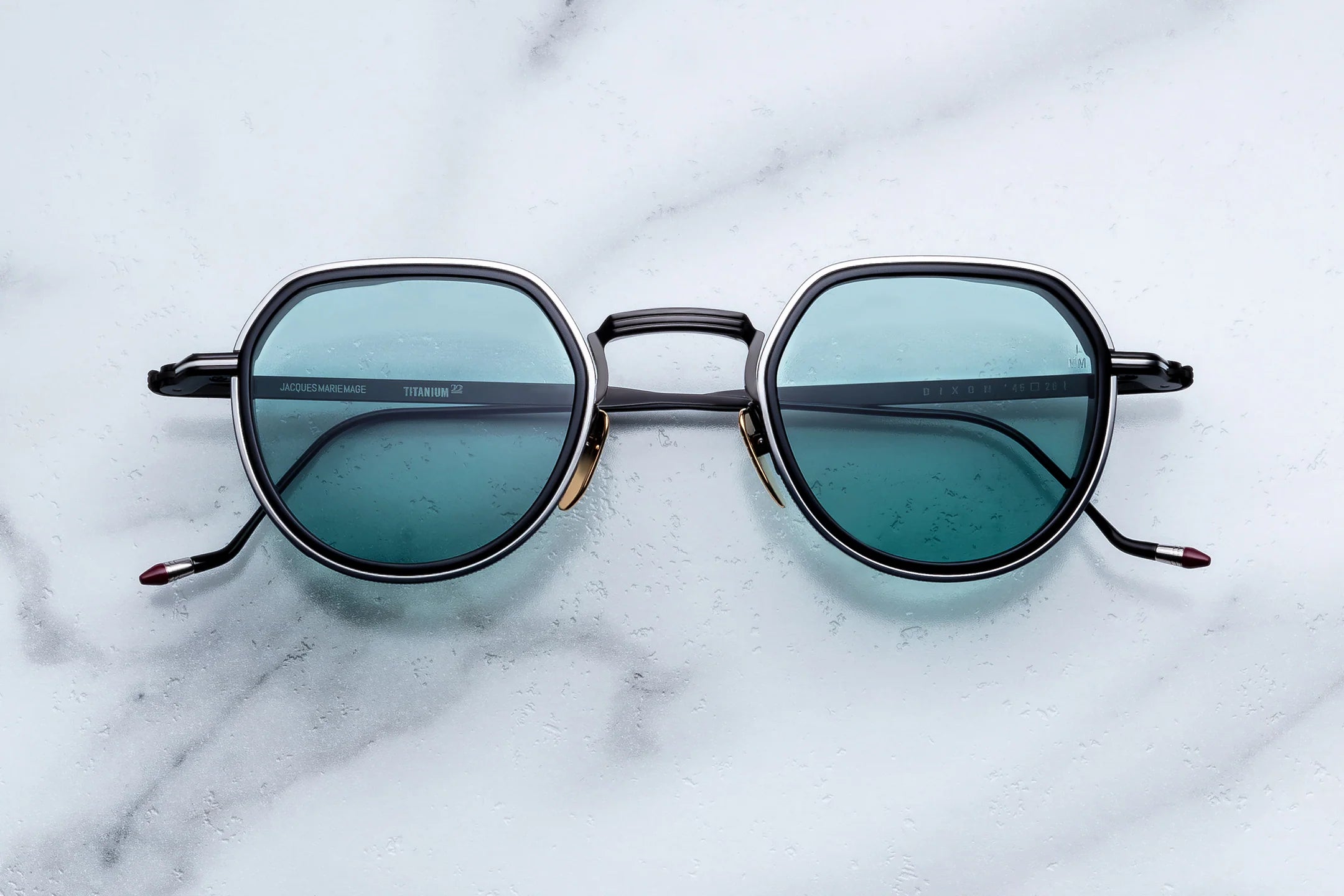 Oversized round sunglasses with a dark gray frame and blue-green tinted lenses, resting on a white marble surface.