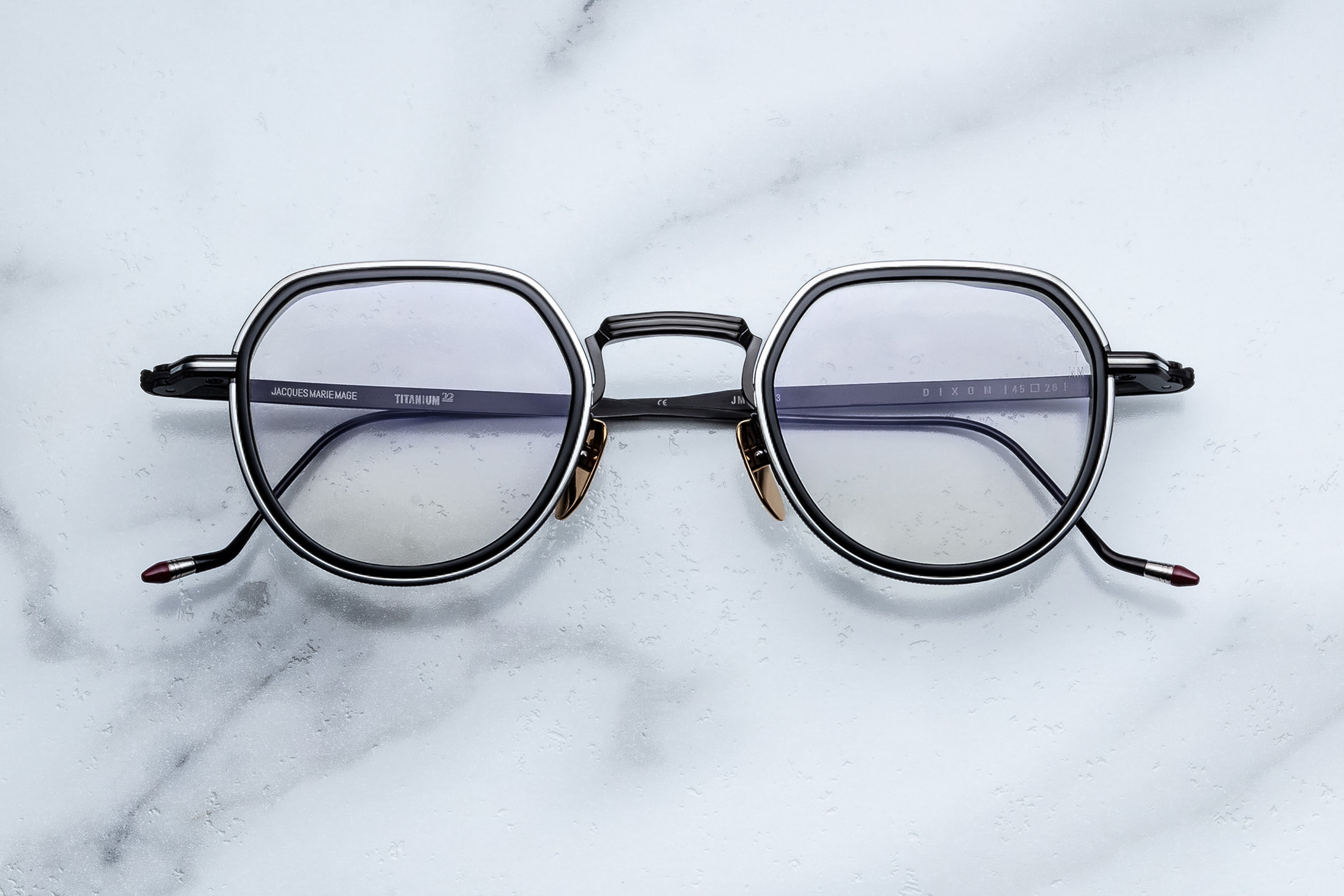 Jacques Marie Mage Dixon eyeglasses by Jacques Marie Mage feature thin black and silver hexagonal frames with clear lenses, elegantly displayed on a white marble surface—an ideal choice for rogue philosophers with an outlaw spirit.