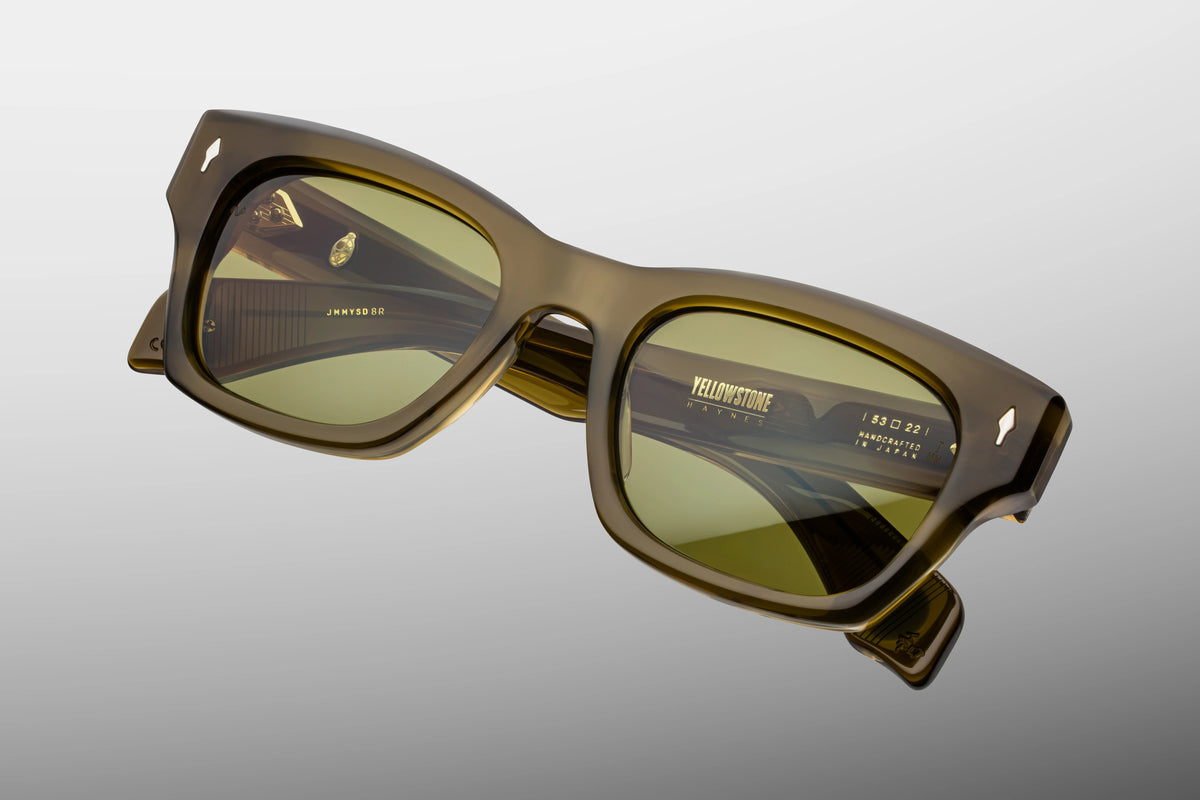 Jacques Marie Mage Yellowstone VII: Haynes In Green Moss ’60s-inspired olive green rectangular sunglasses with gold accents, yellow-tinted lenses, and Yellowstone Haynes printed on the right lens.