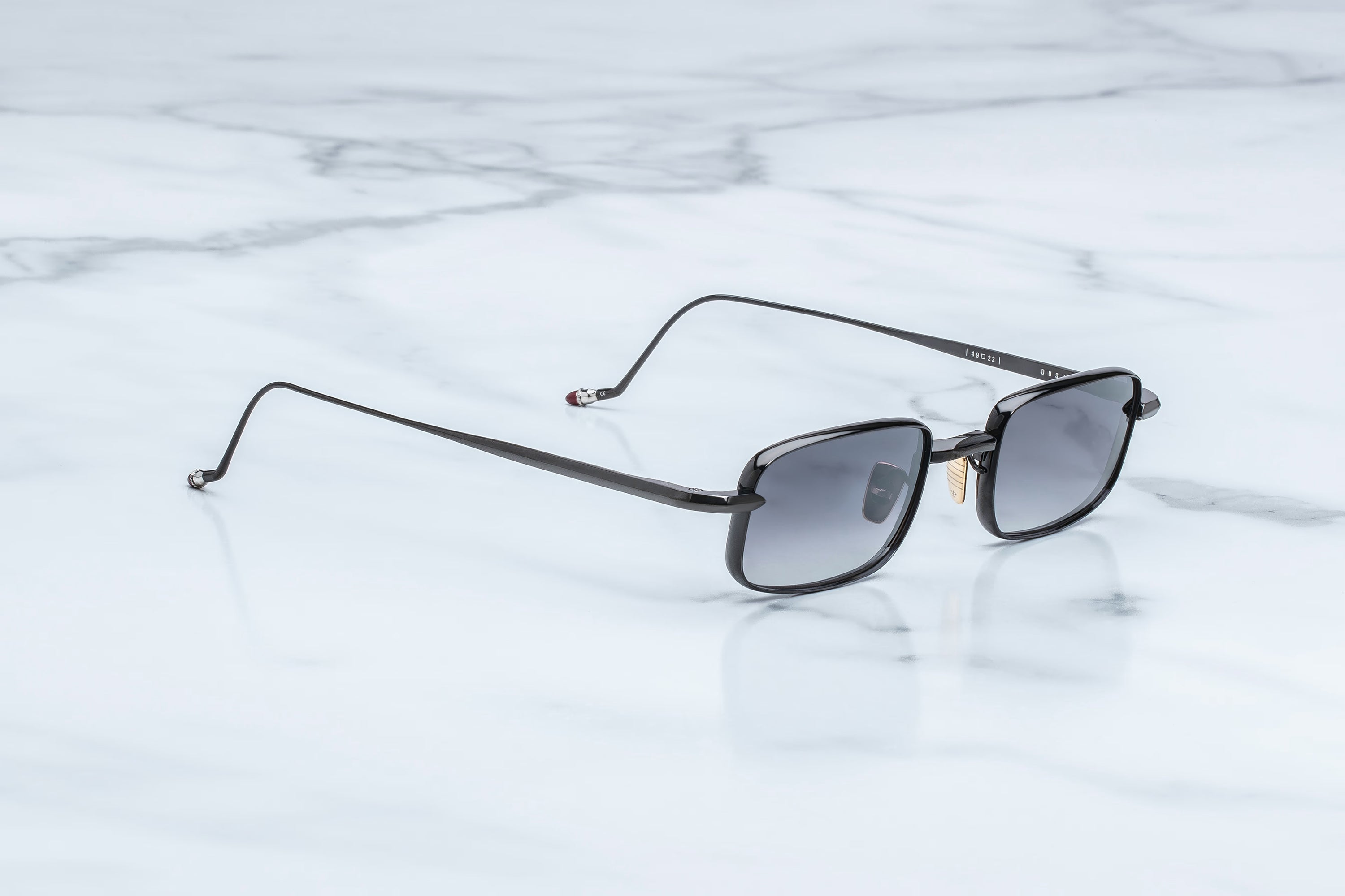 A pair of slim black metal sunglasses with gradient gray lenses, resting on a white marble surface.