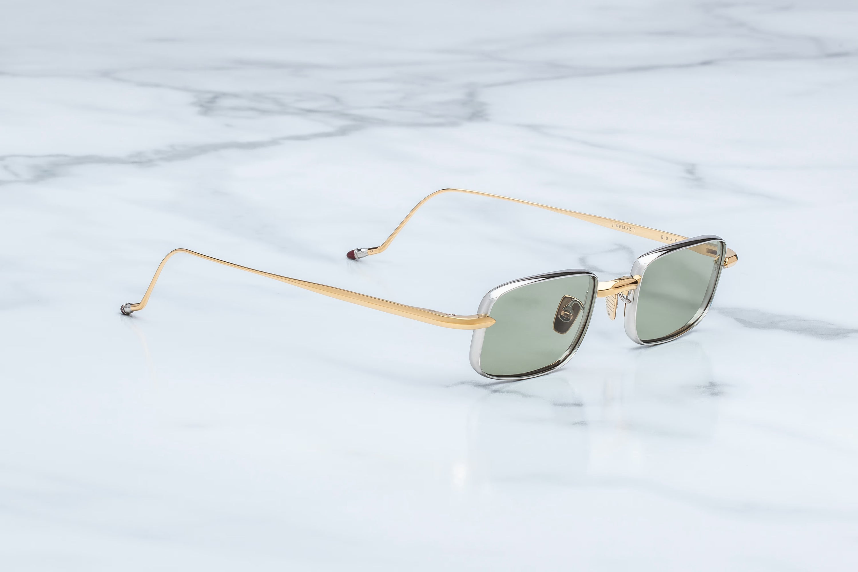 A pair of square-frame sunglasses with gold metal rims, gold temples, and muted green lenses, resting on a white marble surface.