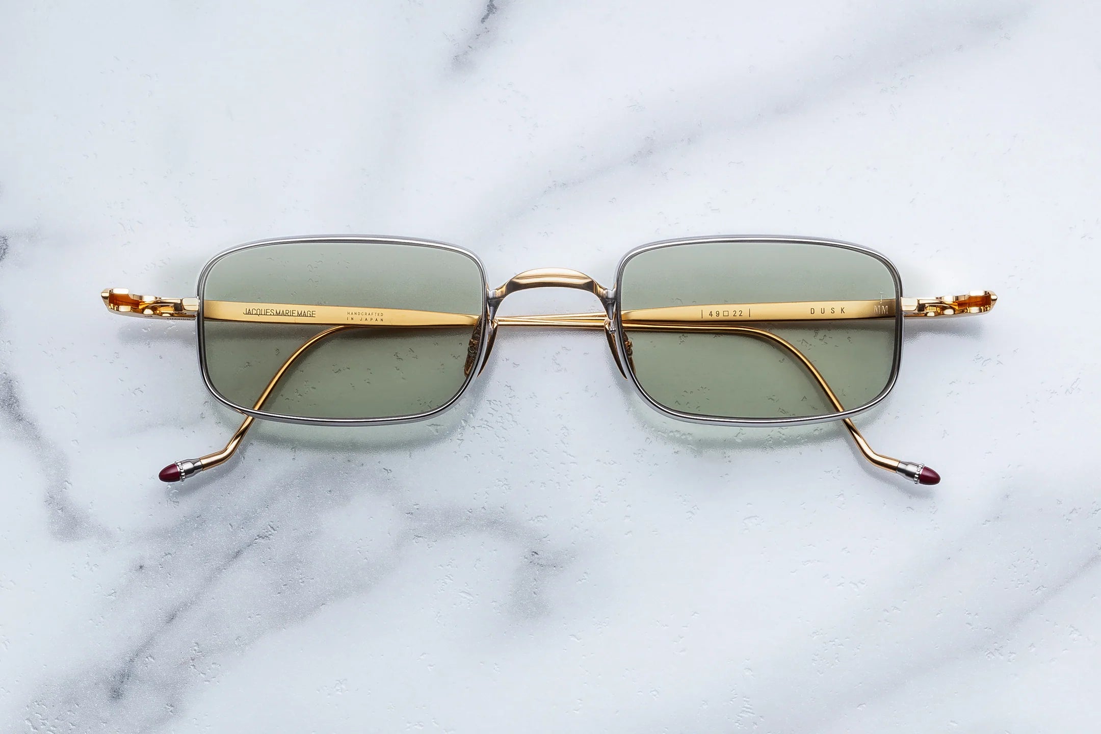 A pair of vintage-style square eyeglasses with gold metal frames, olive green tinted lenses, and decorative gold-toned temples, displayed flat on a white marble surface.