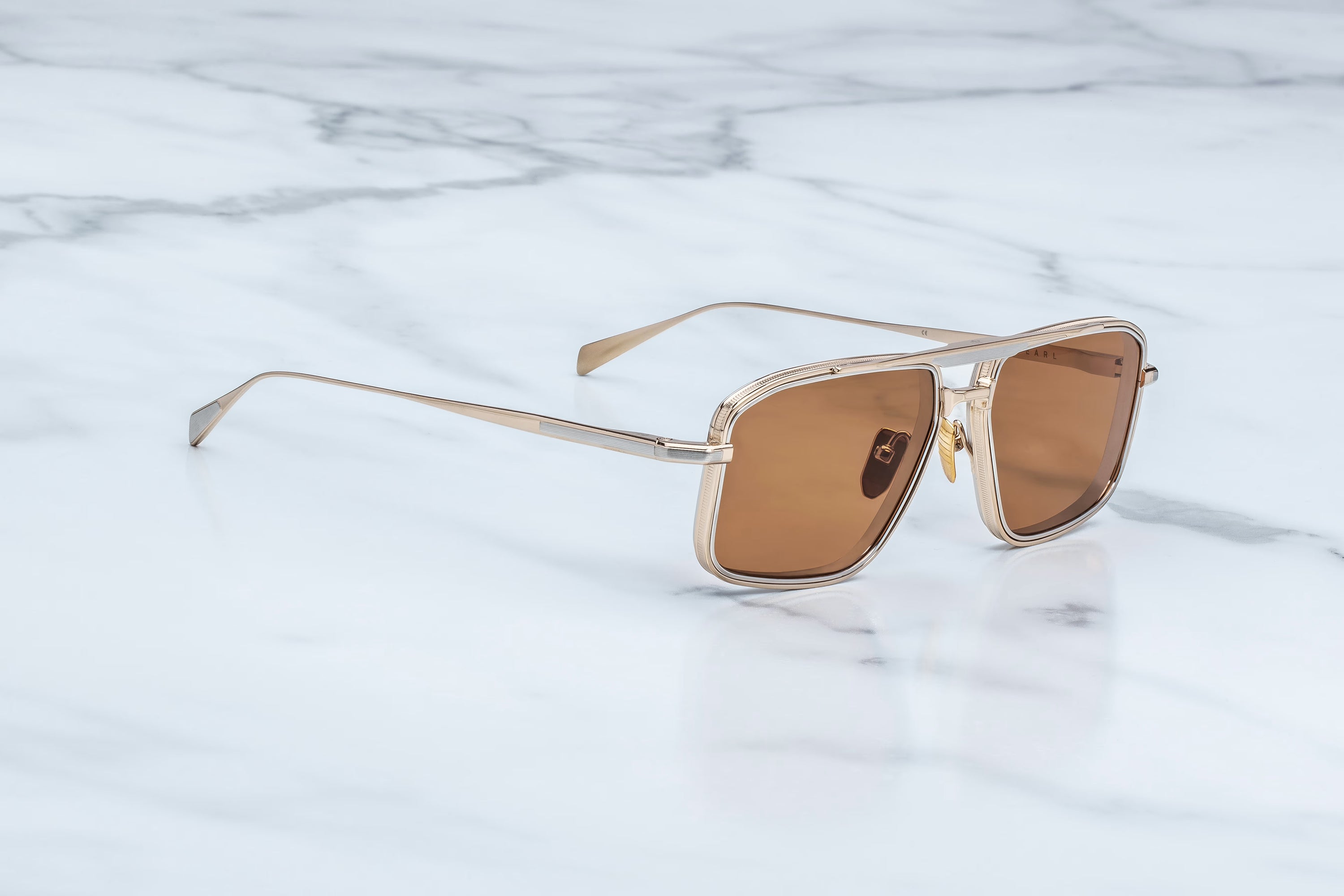 A pair of square aviator-style sunglasses with a thin gold metal frame and warm brown tinted lenses, resting on a white marble surface.