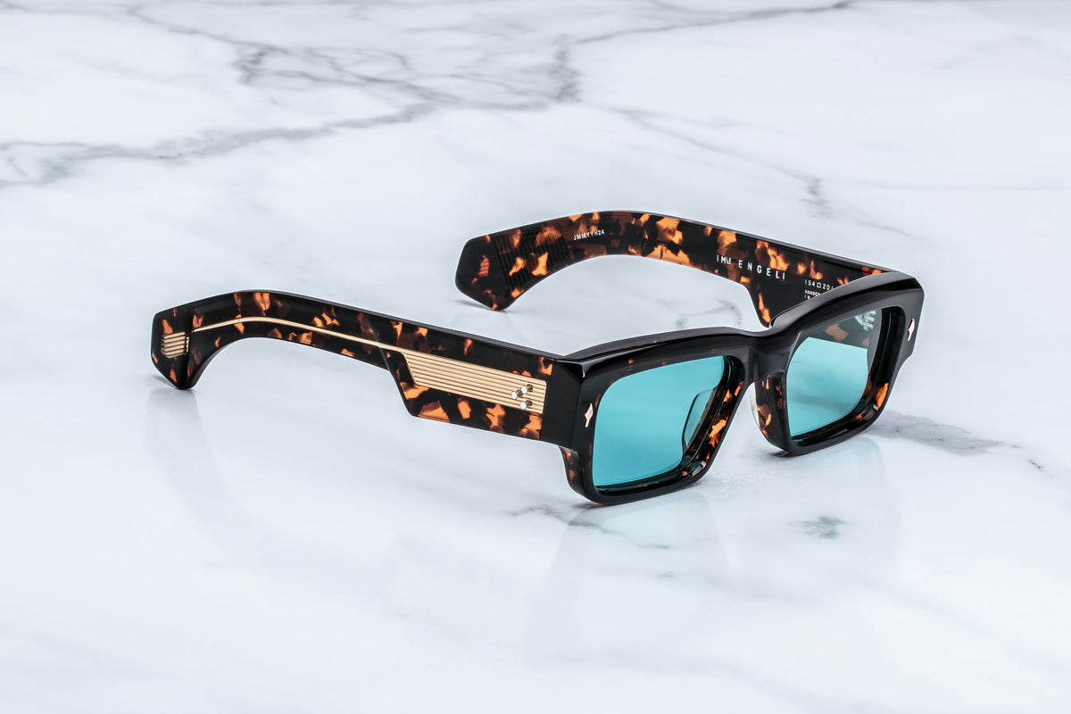 Jacques Marie Mage Engel sunglasses by Jacques Marie Mage, featuring a rectangular silhouette and thick tortoiseshell frames, handcrafted in Japan with blue-tinted lenses, rest on a white marble surface.