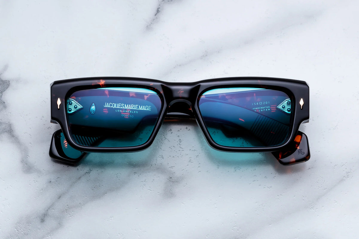 Jacques Marie Mage Engel sunglasses in black, featuring a ’60s-inspired rectangular silhouette and blue-tinted lenses, are placed on white marble. Handcrafted in Japan as a limited-edition piece, these Jacques Marie Mage Engel spectacles have lenses that reflect text and images, including the brand name.