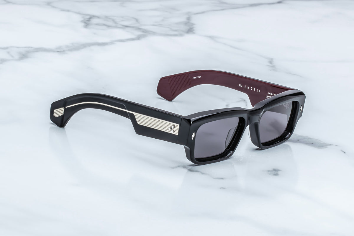 Jacques Marie Mage Engel sunglasses by Jacques Marie Mage feature a bold black rectangular silhouette, dark lenses, silver striped accents on the temples, and maroon-hued inner arms. These limited-edition eyewear pieces are meticulously handcrafted in Japan and showcased on a white marble surface.