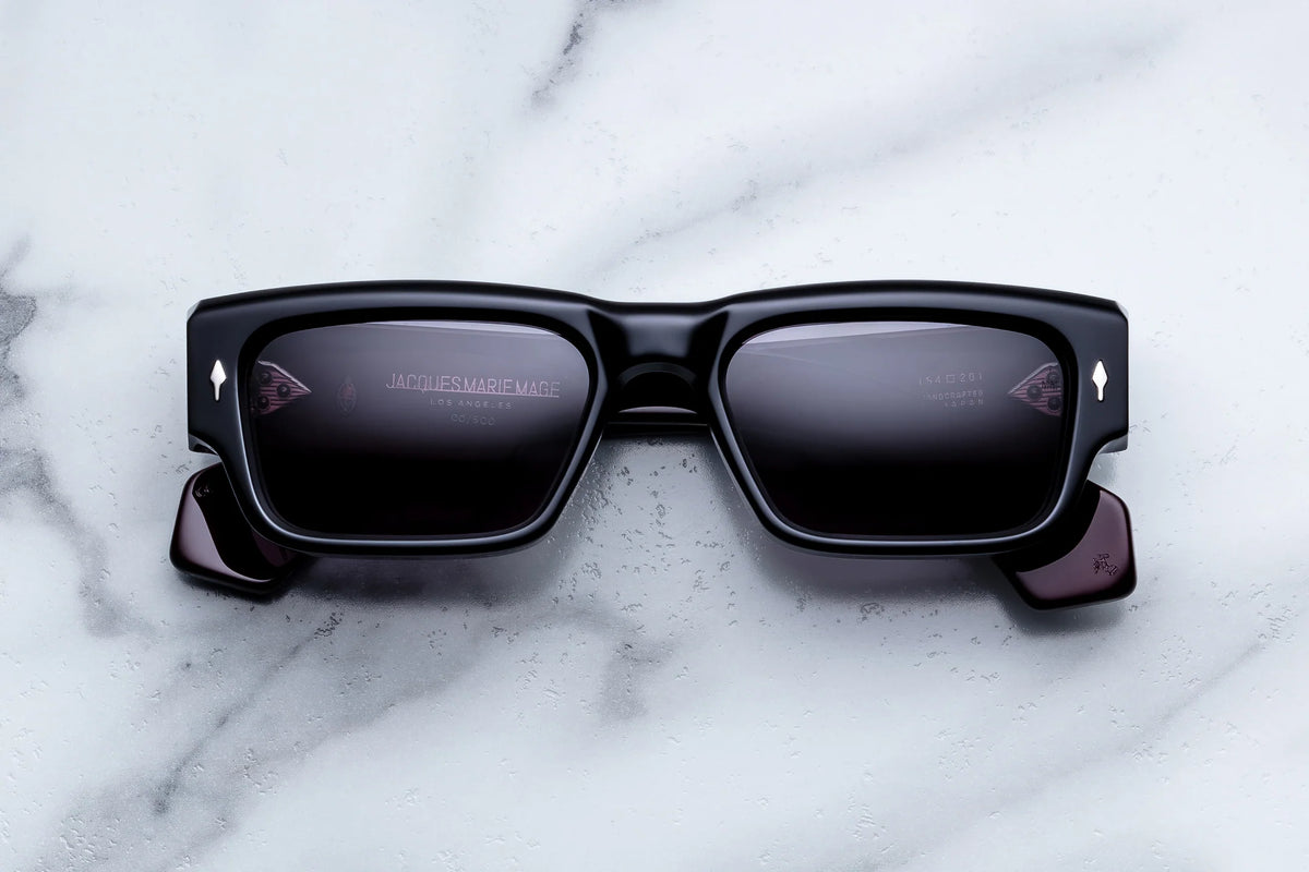 Jacques Marie Mage Engel sunglasses by Jacques Marie Mage rest on a white marble surface. Handcrafted in Japan as a limited-edition release, these black rectangular spectacles feature a ’60s-inspired silhouette, thick frames, dark reflective lenses, and small diamond-shaped accents near the hinges.
