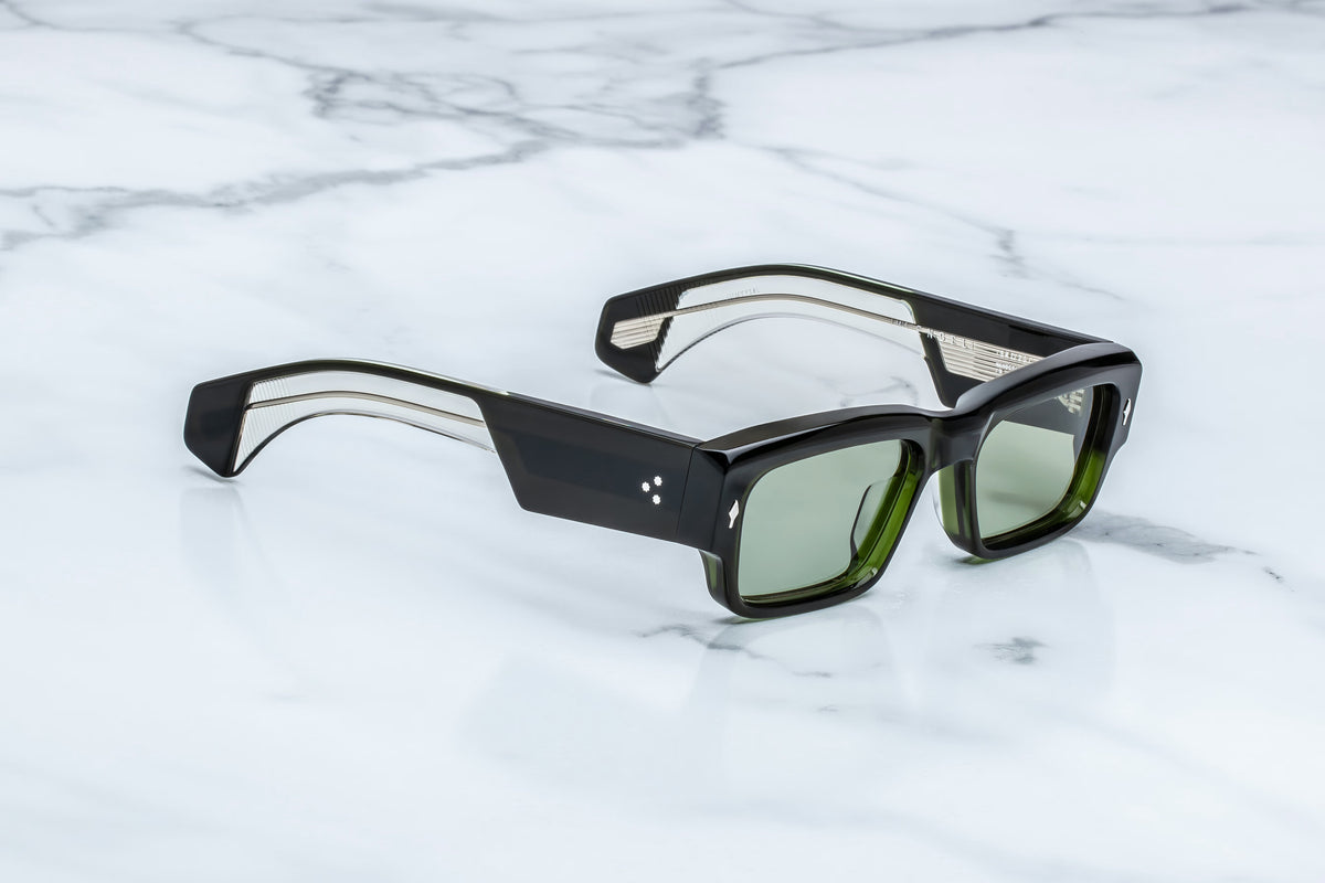 Jacques Marie Mage Engel sunglasses by Jacques Marie Mage, featuring a bold rectangular silhouette with green-tinted lenses and thick black and clear handcrafted plastic frames made in Japan, elegantly displayed on a white marble surface.