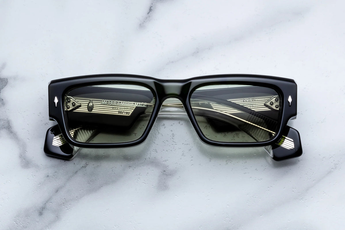 The Jacques Marie Mage Engel eyeglasses by Jacques Marie Mage are showcased on a white marble surface. These black, thick-framed rectangular spectacles feature reflective lenses with a metallic, striped pattern. Expertly handcrafted in Japan, the limited-edition frames display subtle silver diamond-shaped accents near the hinges and embody a refined ’60s-inspired silhouette that skillfully merges vintage charm with contemporary craftsmanship.