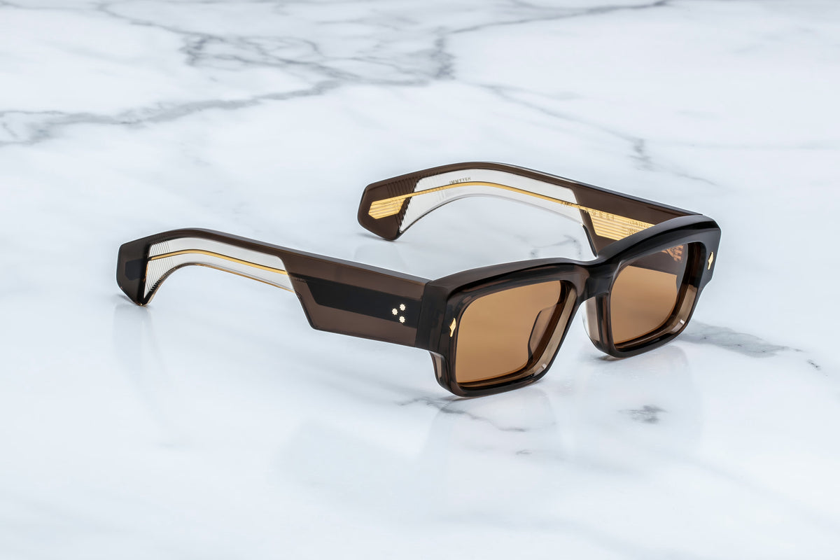 Jacques Marie Mage Engel sunglasses by Jacques Marie Mage, featuring a refined rectangular silhouette in brown with a thick frame and gold accents, rest on a white marble surface—handcrafted in Japan as true limited-edition spectacles.