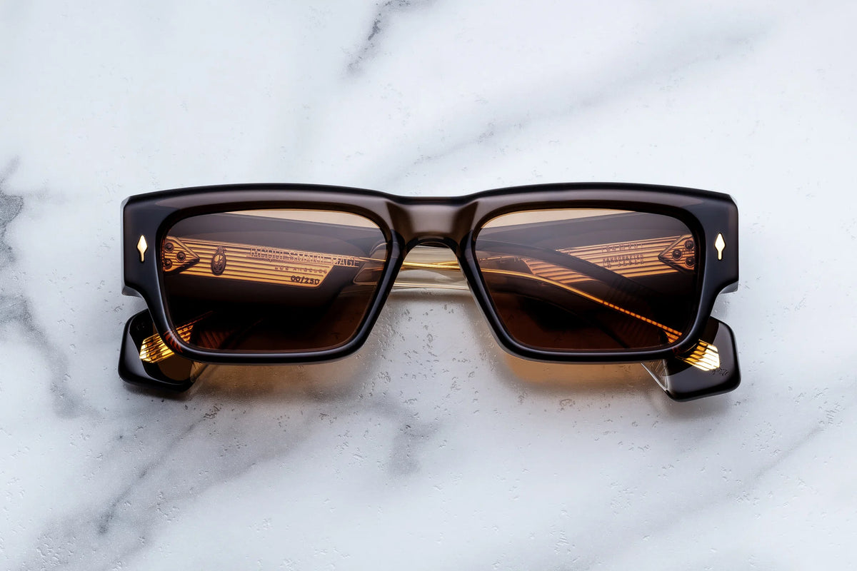 Introducing the Jacques Marie Mage Engel by Jacques Marie Mage—limited-edition rectangular black sunglasses handcrafted in Japan, featuring brown tinted lenses and elegant gold accents. With a ’60s-inspired silhouette, these spectacles showcase thick frames adorned with subtle decorative details at the corners. Pictured resting atop a white marble surface.