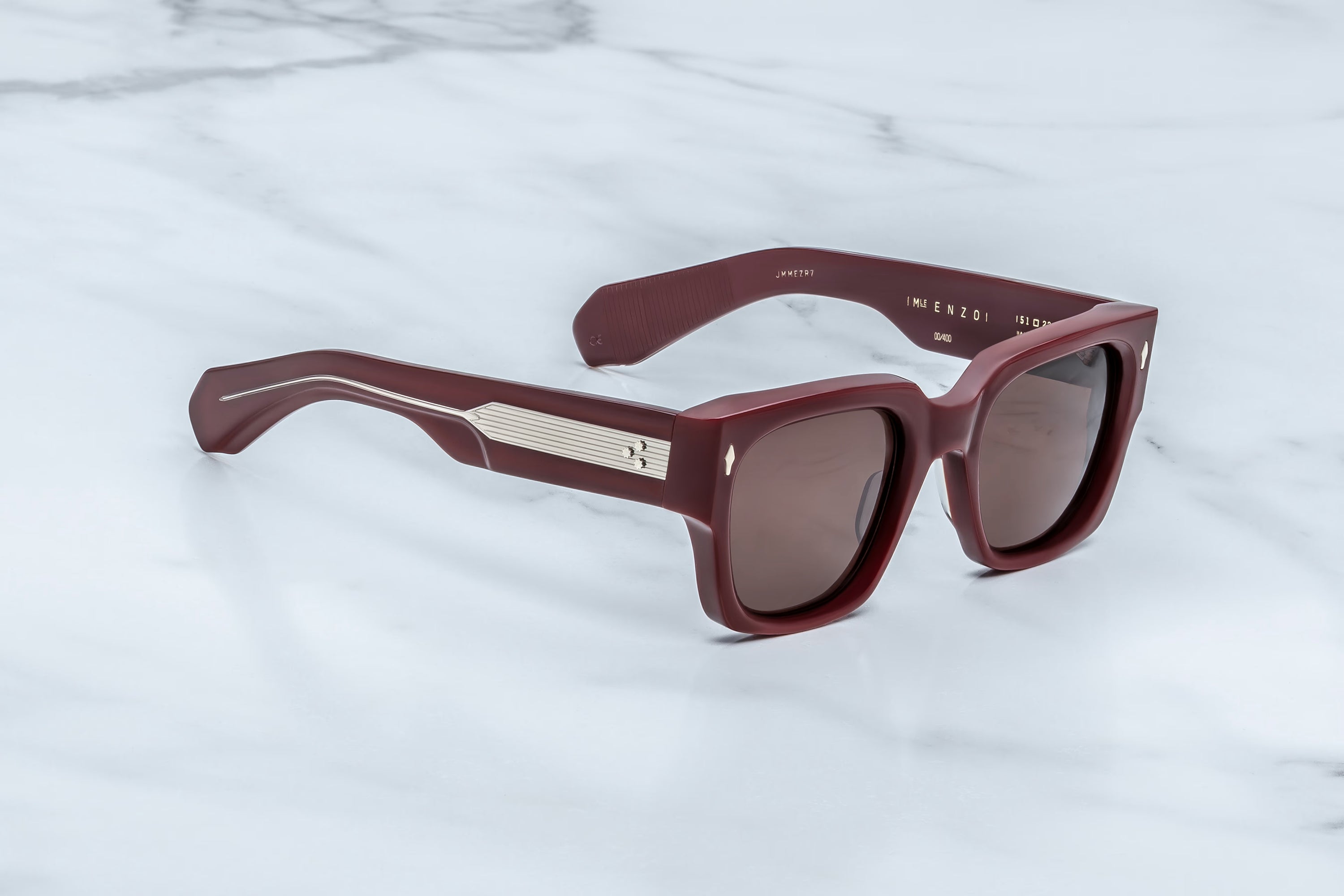 A pair of square-frame sunglasses with a translucent burgundy frame and dark brown lenses, resting on a white marble surface. 