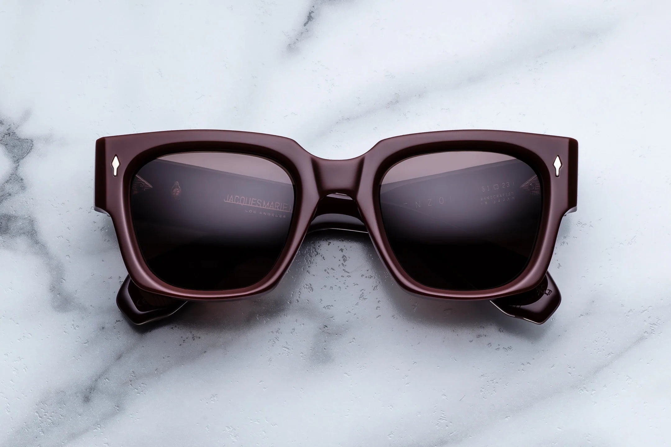 The Jacques Marie Mage Enzo, dark brown square sunglasses with black lenses inspired by iconic frames reminiscent of Enzo Ferrari’s style, are placed on a white marble surface.