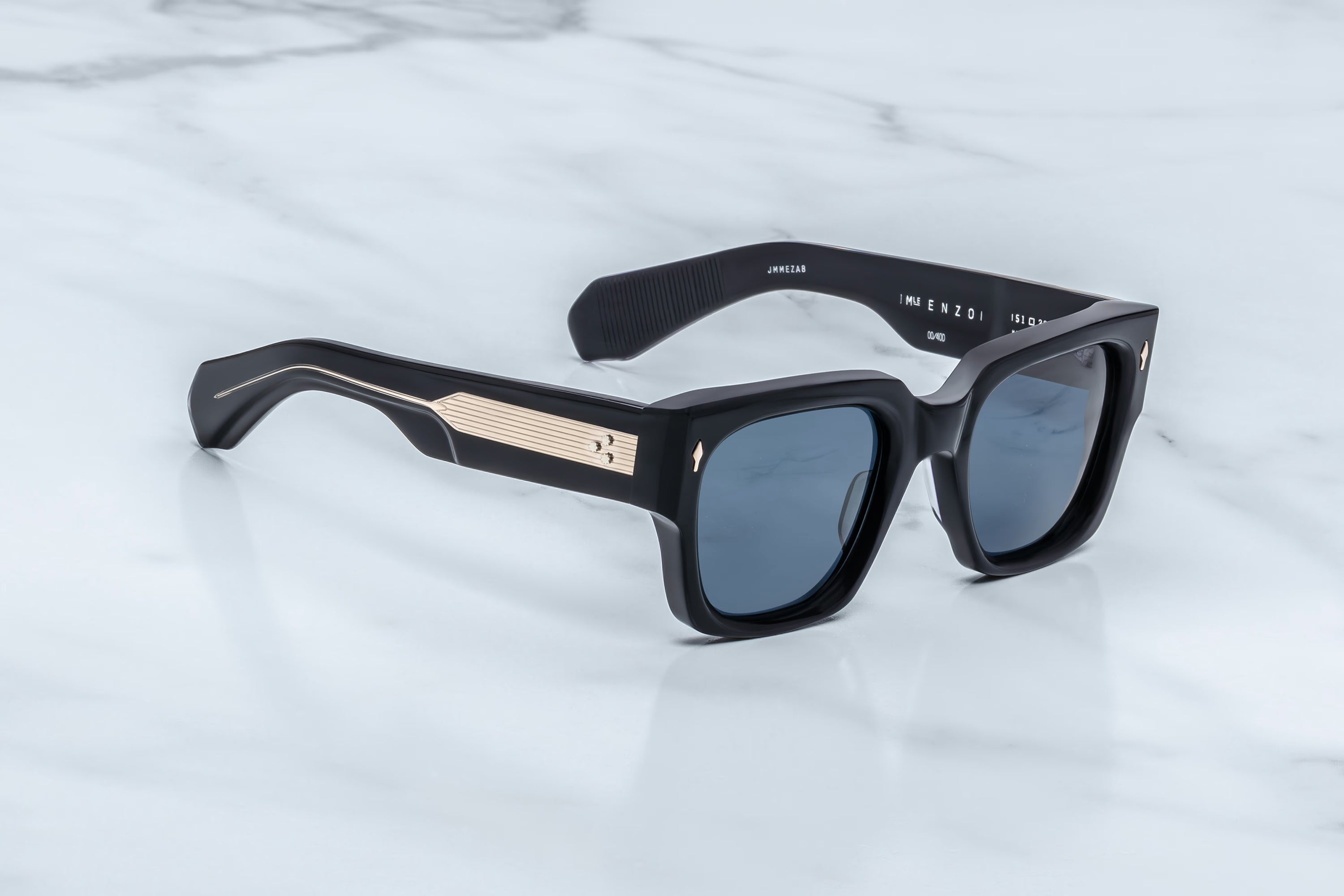 The Jacques Marie Mage Enzo sunglasses by Jacques Marie Mage—featuring navy square frames, dark blue lenses, and gold detailing on the arms—rest atop a white marble surface, capturing the timeless allure of iconic eyewear inspired by Enzo Ferrari.
