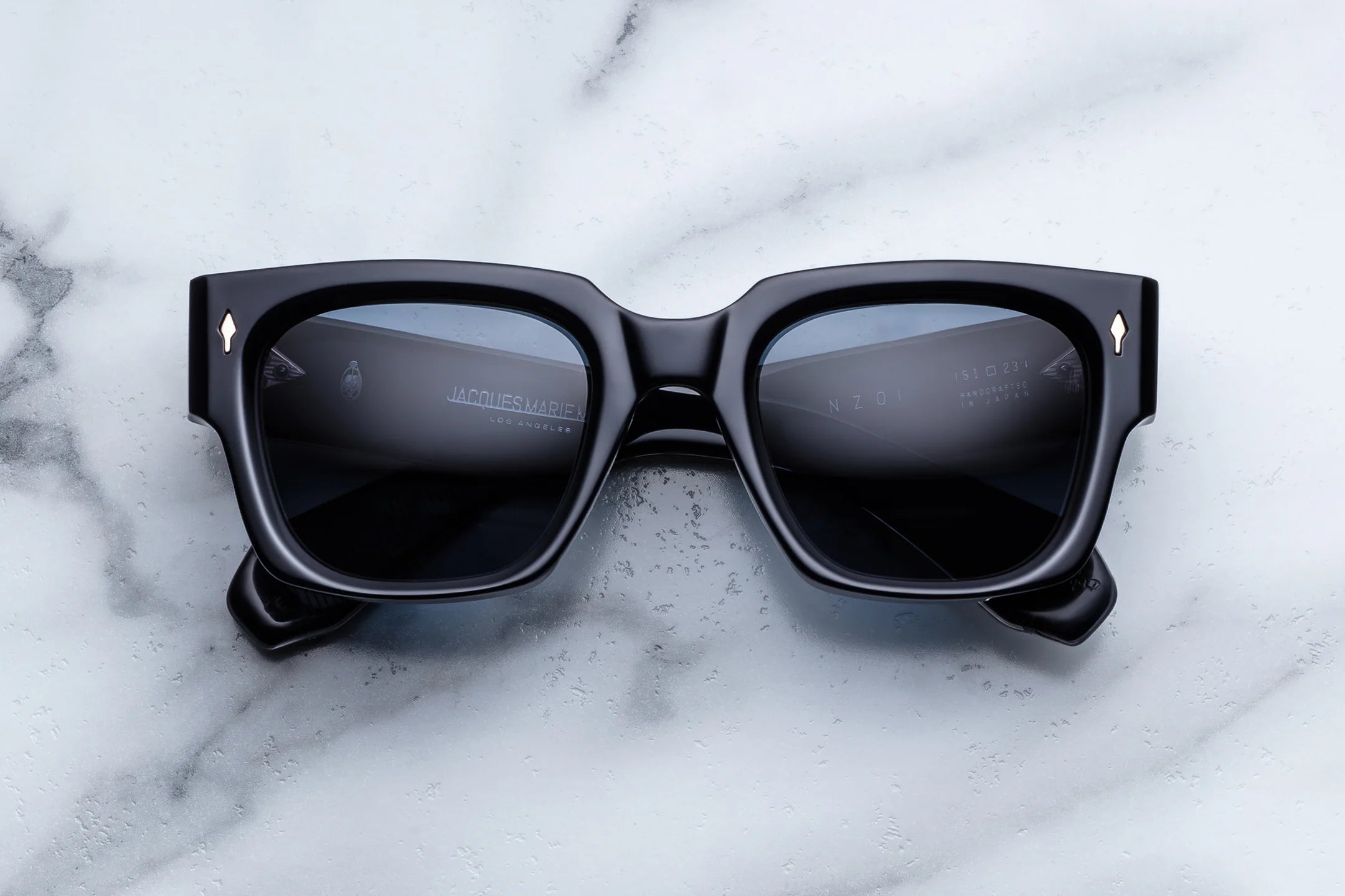 Jacques Marie Mage Enzo navy square-frame sunglasses with dark lenses rest on a white marble surface. Crafted by Jacques Marie Mage, these iconic frames feature thick construction and small metallic accents near the hinges, evoking the timeless style of limited-edition glasses inspired by Enzo Ferrari.