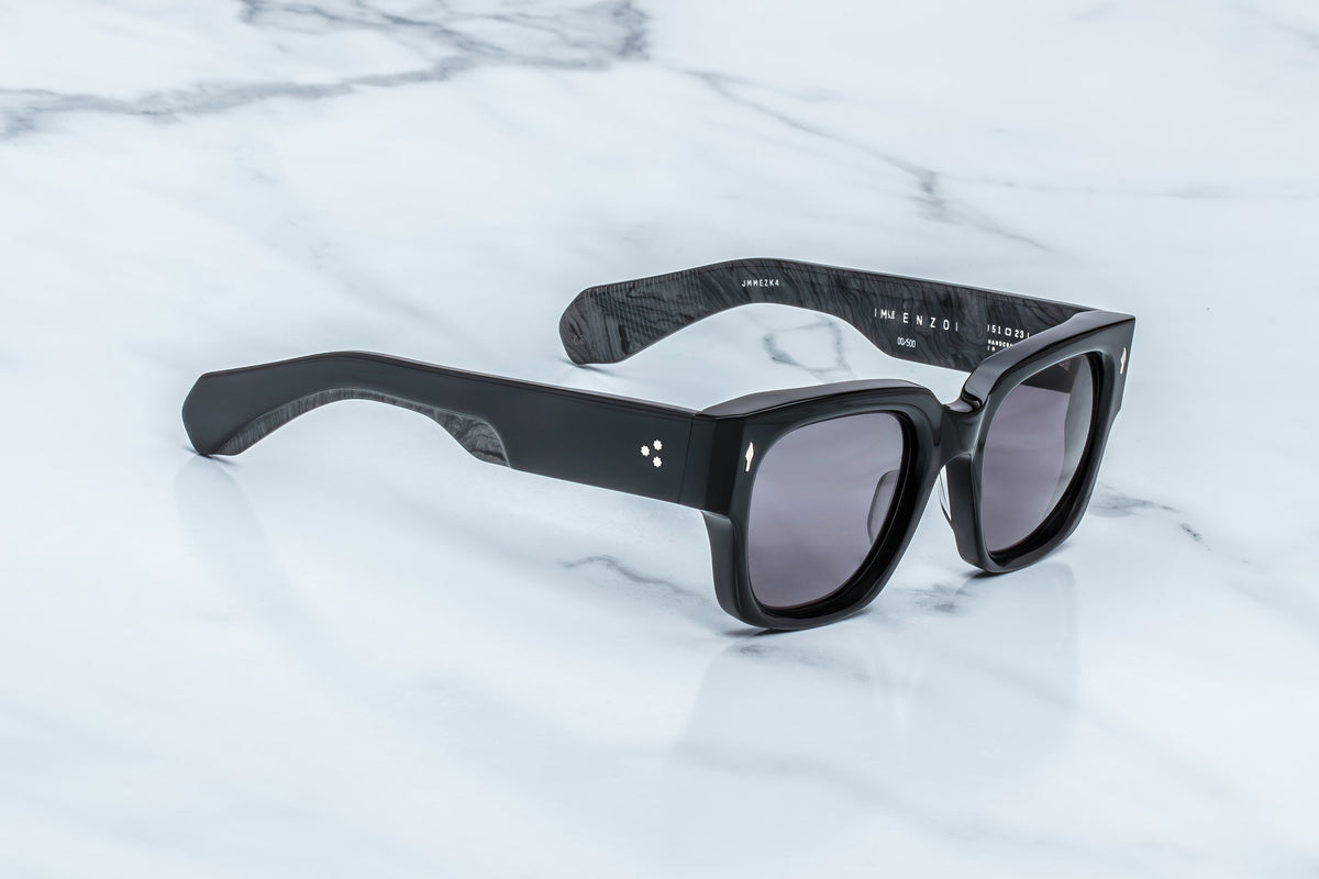 Jacques Marie Mage Enzo sunglasses by Jacques Marie Mage, featuring a black rectangular frame with dark lenses, are resting on a white marble surface. The thick temples display three small white dots on the hinge area of one arm—an understated detail reminiscent of iconic frames like the original Enzo Ferrari glasses.