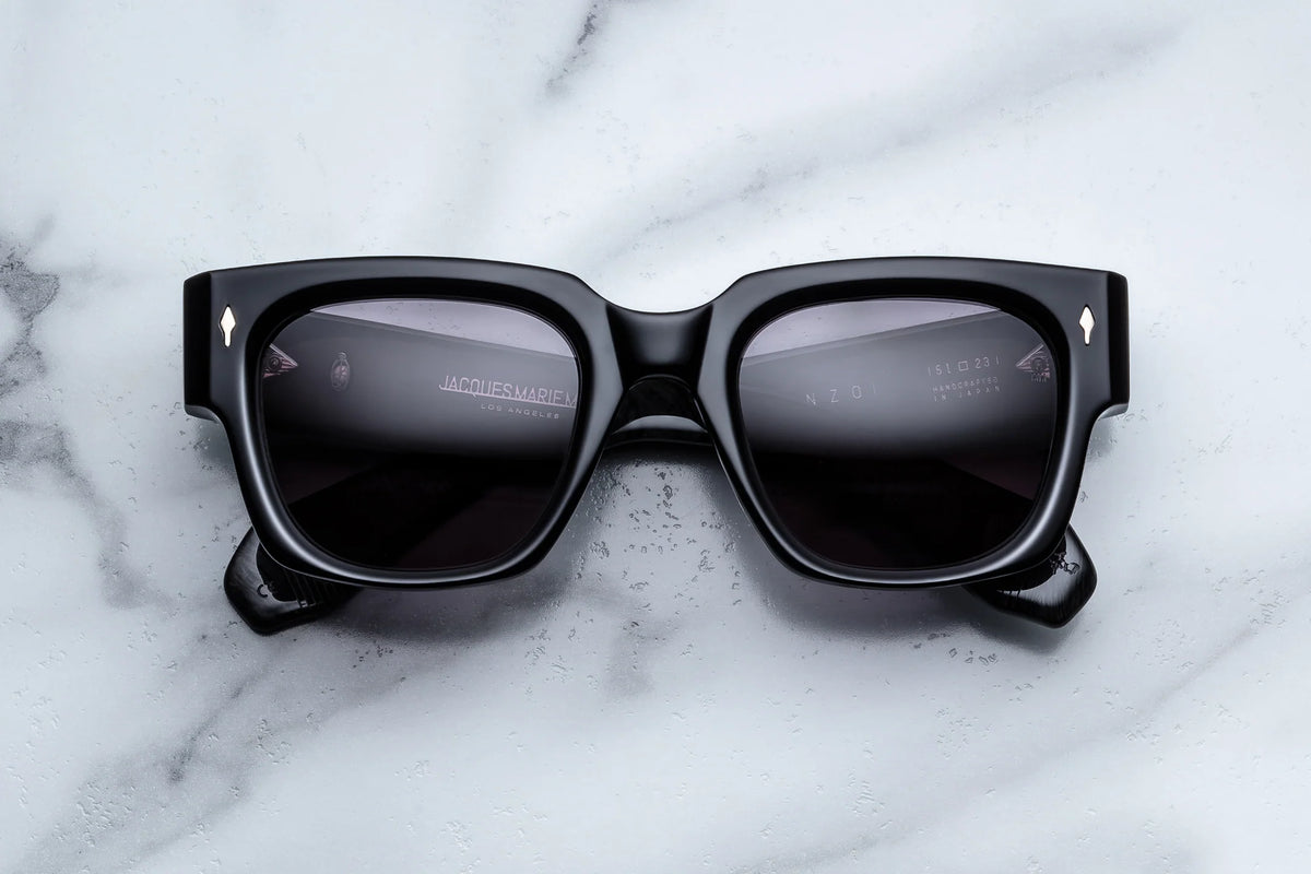 Jacques Marie Mage Enzo sunglasses by Jacques Marie Mage—featuring black, square-shaped frames with dark lenses and small silver diamond-shaped accents at the upper corners, reminiscent of iconic Enzo Ferrari styles—are elegantly displayed on a white marble surface.